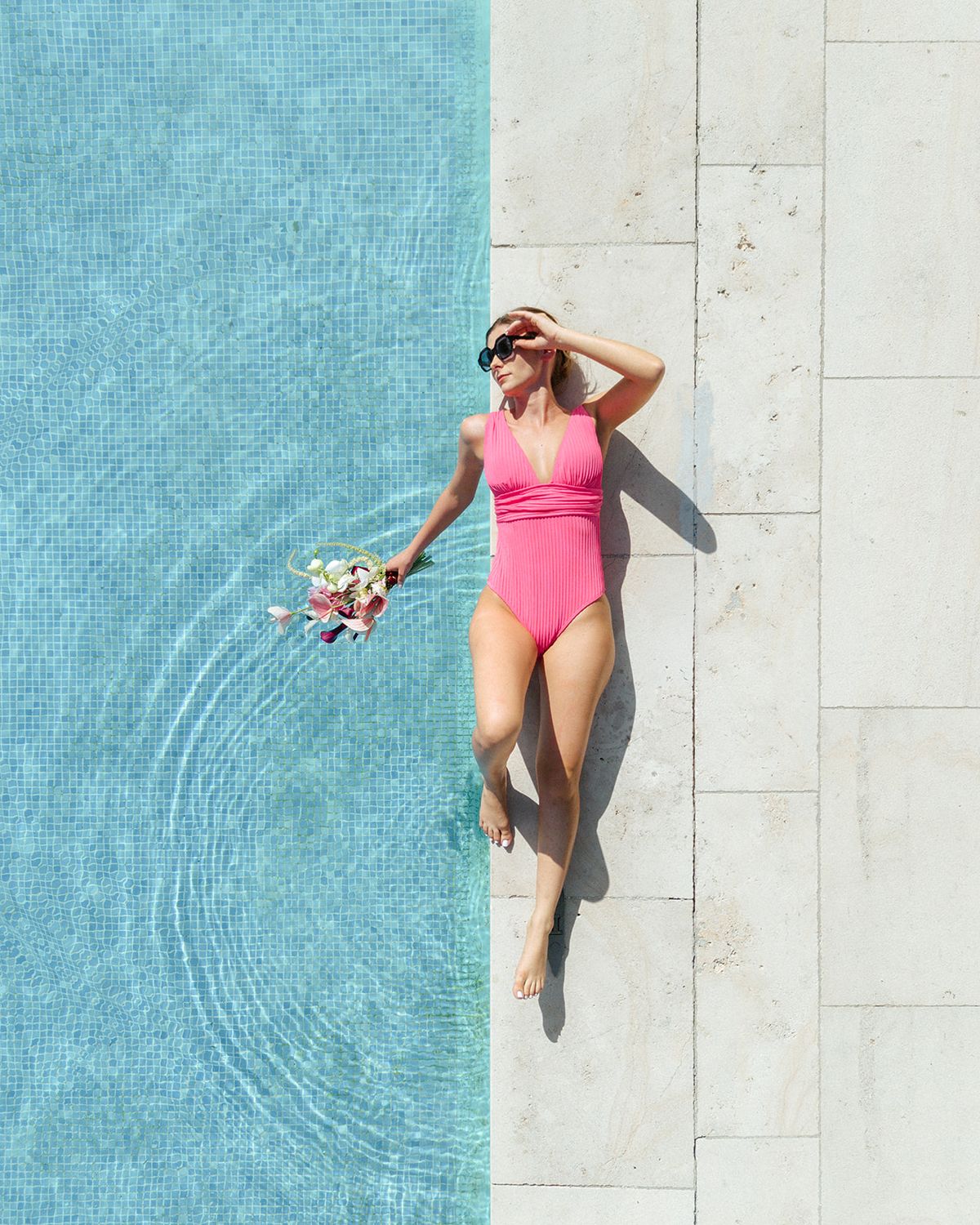 Overhead view of the bride in a pink swimsuit at the pool edge holding a small posy