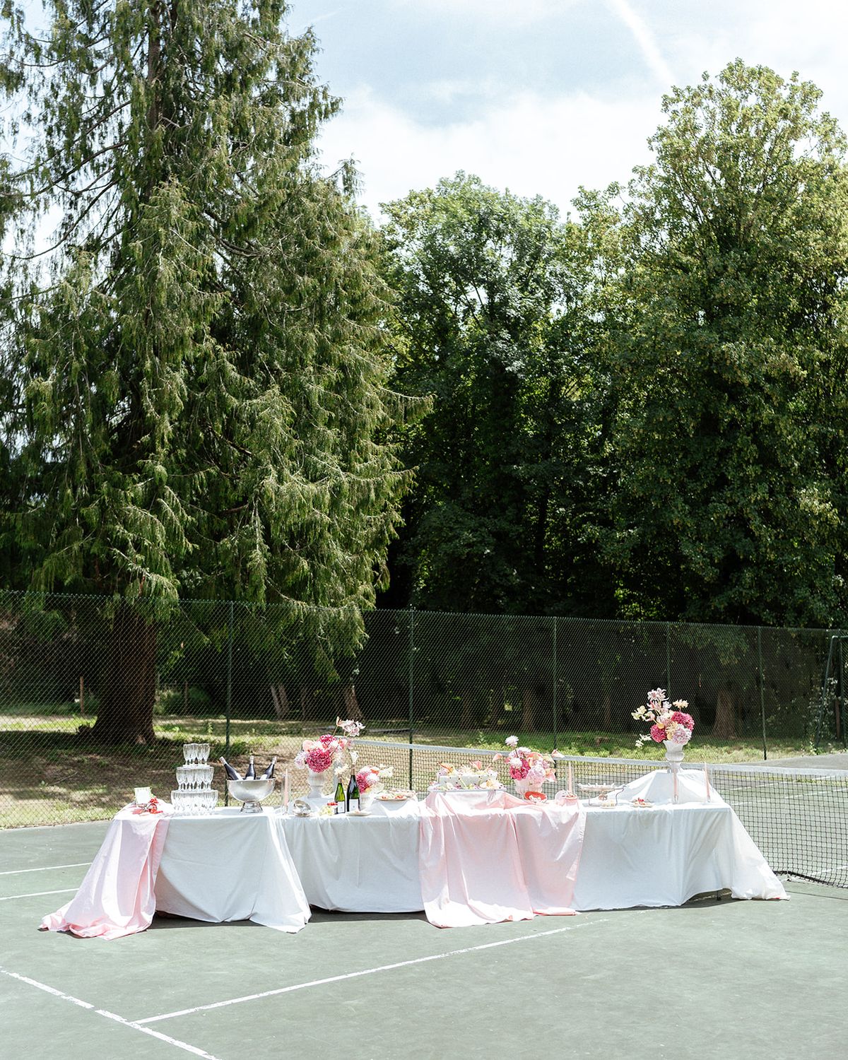 Tables draped in white and pink set on a tennis court in front of tall conifers