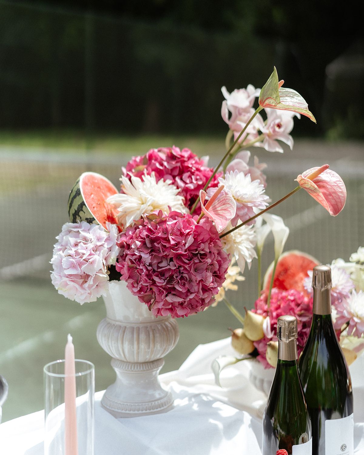 Tall urn of pink hydrangea and anthurium on a white table with champagne and candles