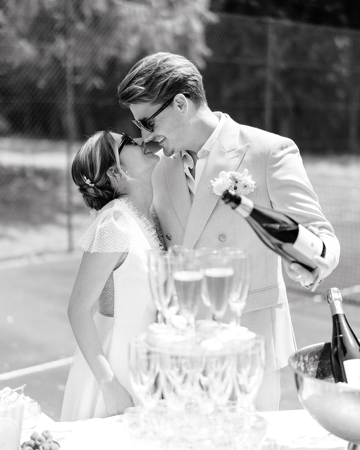 Couple in sunglasses beside a champagne coupe tower on the tennis court, the groom pouring