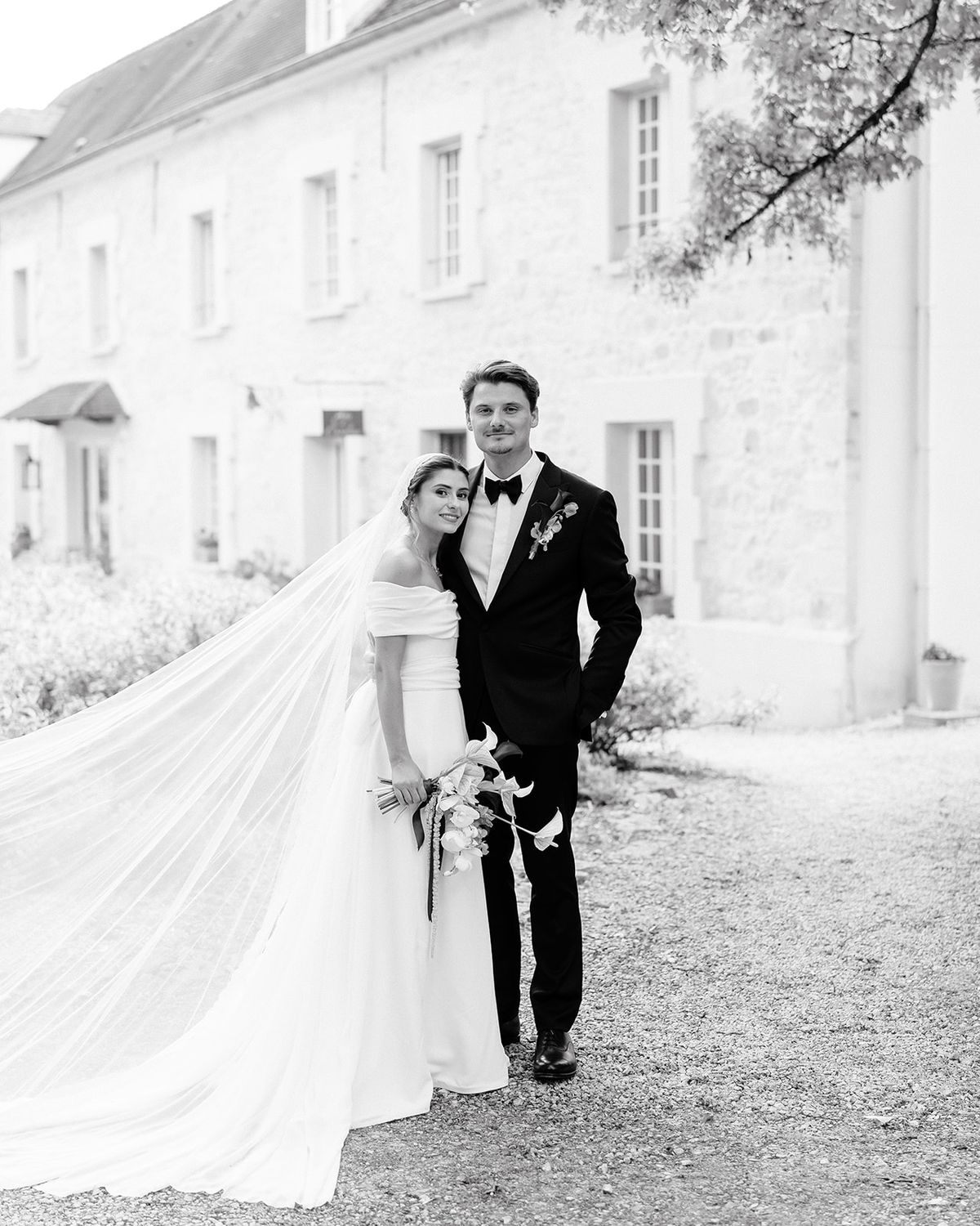 Portrait of the couple walking in front of the stone house with the veil trailing behind