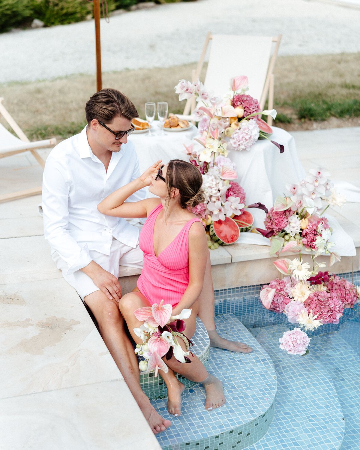 Couple sitting at the pool edge with champagne and pastries on a draped table