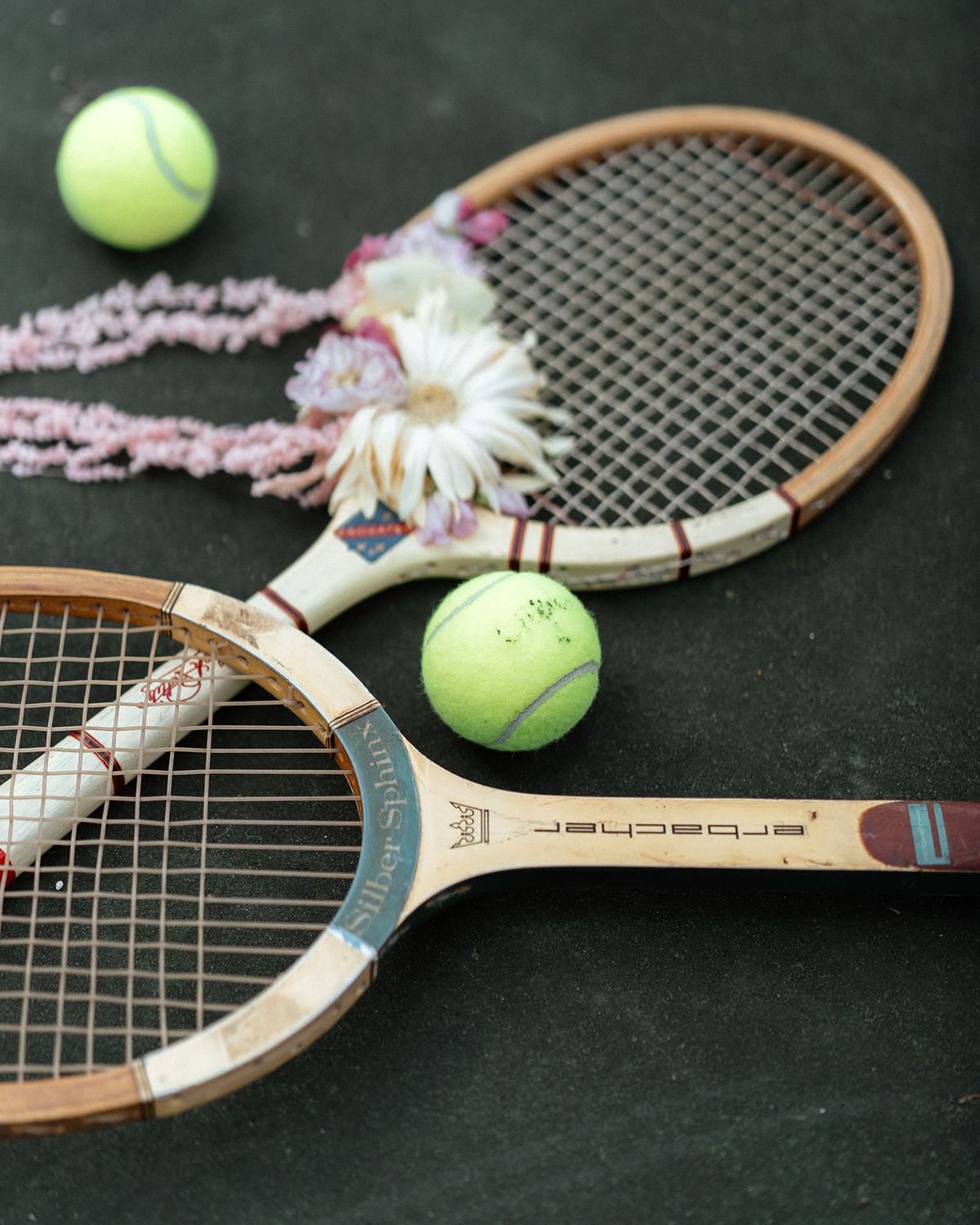 Two vintage wooden tennis rackets and three tennis balls arranged with dahlias on a green court