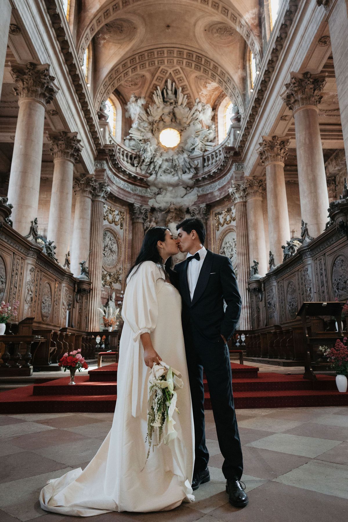 A bride and groom share a kiss at the altar of a grand Baroque-style church, standing on a tiled stone floor with a deep red carpet runner leading toward the ornate apse behind them. The bride wears an ivory long-sleeve dress with voluminous bishop sleeves and a flowing train, holding a loose trailing bouquet of white calla lilies and delicate greenery; the groom wears a black tuxedo with a black bow tie. The church interior features tall fluted columns, heavily carved wooden choir stalls lining both sides, elaborate gilded and sculpted stucco figures surrounding the altar, and a domed ceiling with decorative coffering. Small arrangements of red and pink flowers are placed at the base of the columns on either side. The shot is a full-length portrait taken from a low angle, emphasizing the height and architectural depth of the interior.