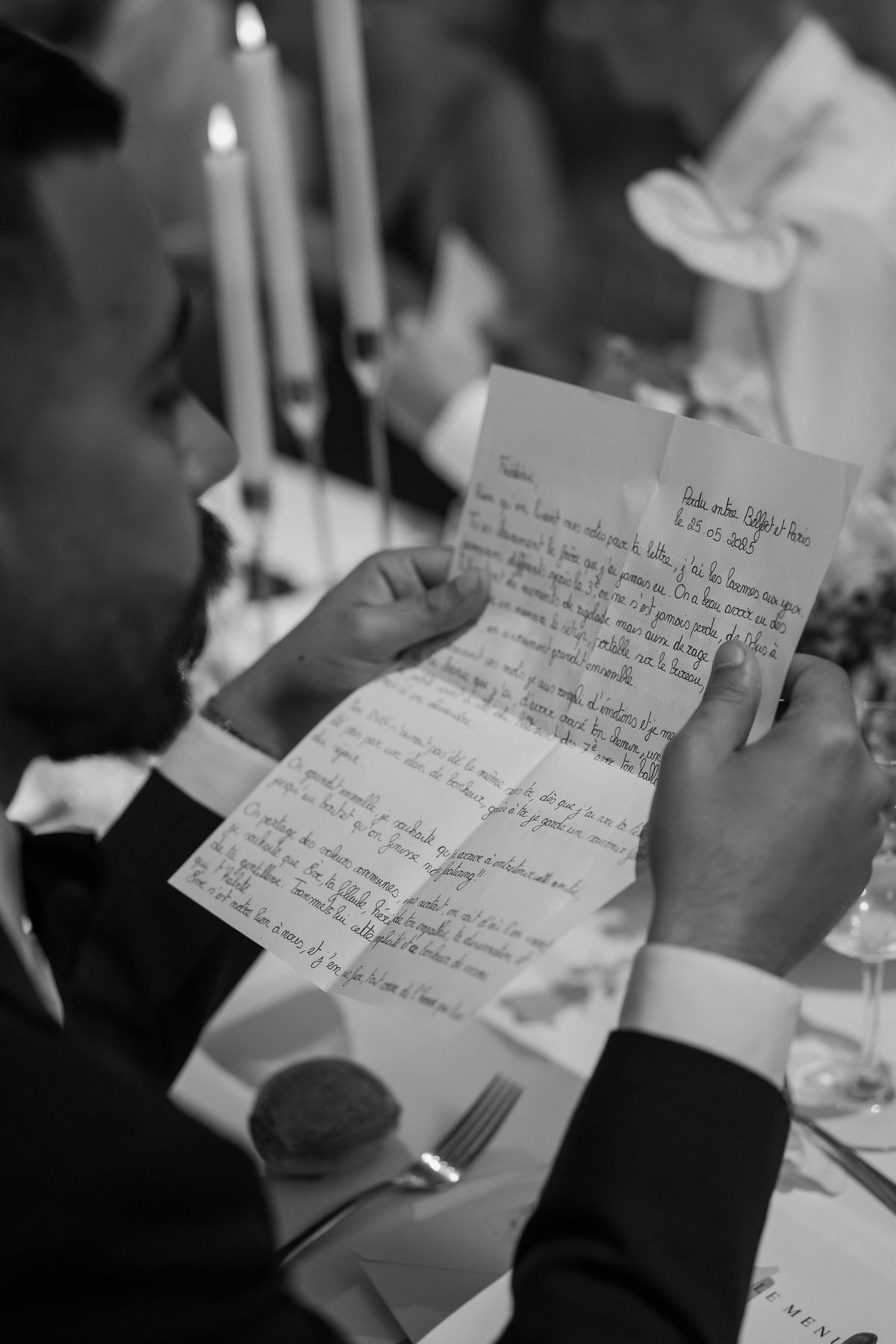 This black-and-white close-up portrait shows a man in a dark suit and white dress shirt reading a handwritten letter during what appears to be a wedding reception dinner. The letter is written in French and dated 25.05.2025, with the header noting 'Perdu entre Belfort et Paris.' The image is shot over his shoulder, with the text of the letter legible in sharp focus against softly blurred background elements including tall taper candles, wine glasses, a fork, and a printed menu card on the white-linened table. The contrast between the dark suit and the bright white paper draws the eye to the letter as the central subject of the frame.