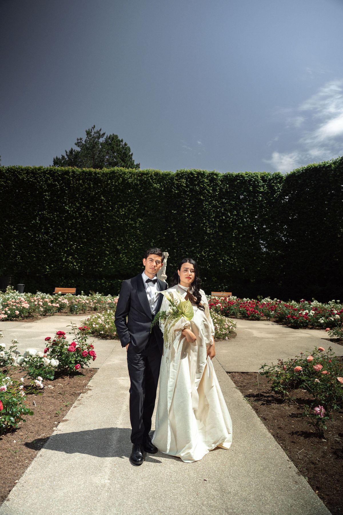 A couple poses for a full-length portrait in a formal outdoor rose garden, standing on a central paved pathway flanked by rose beds in red, pink, and white. The groom wears a black tuxedo with a white dress shirt and black bow tie, while the bride wears a structured ivory gown with voluminous long sleeves and a full skirt; she carries a bouquet featuring anthurium leaves, white calla lilies, and trailing greenery. A white classical statue is visible in the background, along with trimmed tall hedges forming a formal garden enclosure and wooden benches on either side. The composition is a wide shot taken from a low angle, giving prominence to the couple against the open sky.