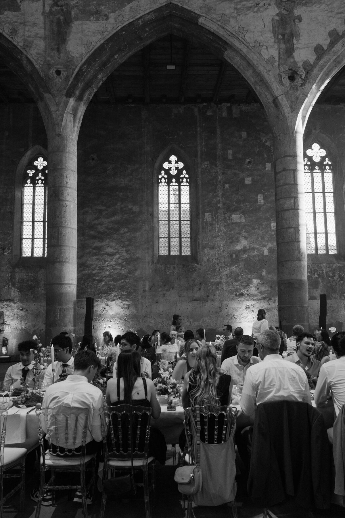 This black-and-white image shows a wedding reception dinner taking place inside a medieval Gothic chapel or abbey, featuring soaring pointed stone arches, tall lancet windows with quatrefoil tracery, exposed stone walls with traces of frescoes, and a wooden beamed ceiling. Warm uplighting illuminates the lower sections of the stone walls, creating strong contrast against the bright windows above. Approximately 30–40 guests are seated at long banquet-style tables arranged along the nave, with Chiavari chairs visible in the foreground; floral centerpieces and candles are visible on the tables. The wide shot captures the full height of the interior architecture while guests dine and converse in the foreground and middle ground. Potential venue feature image.