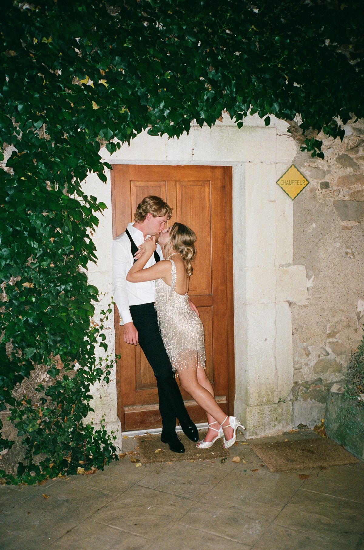 The couple kiss in front of a wooden door set in a stone wall covered with green ivy, the bride in a short sparkling dress.