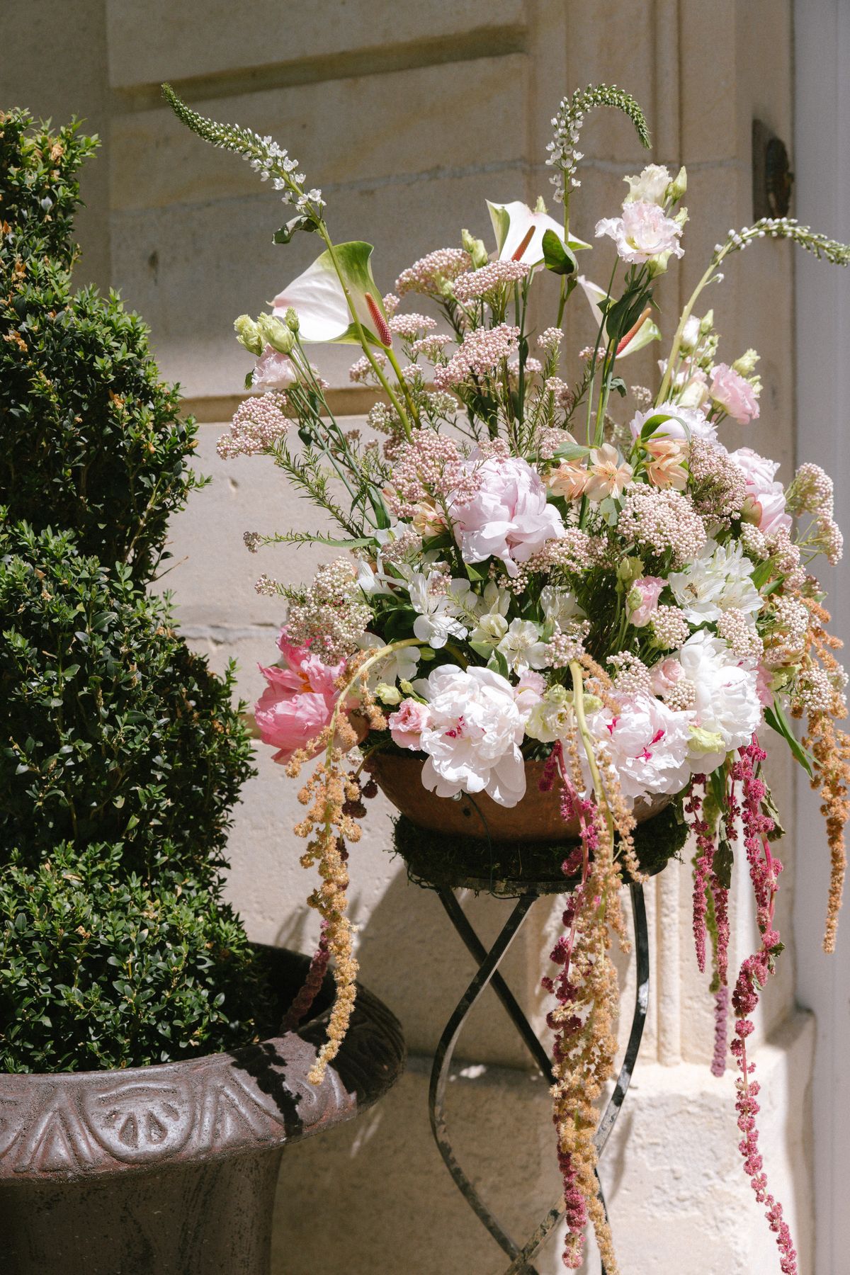 A tall arrangement of pale pink flowers and trailing foliage sits on a dark metal stand beside a pale stone wall.