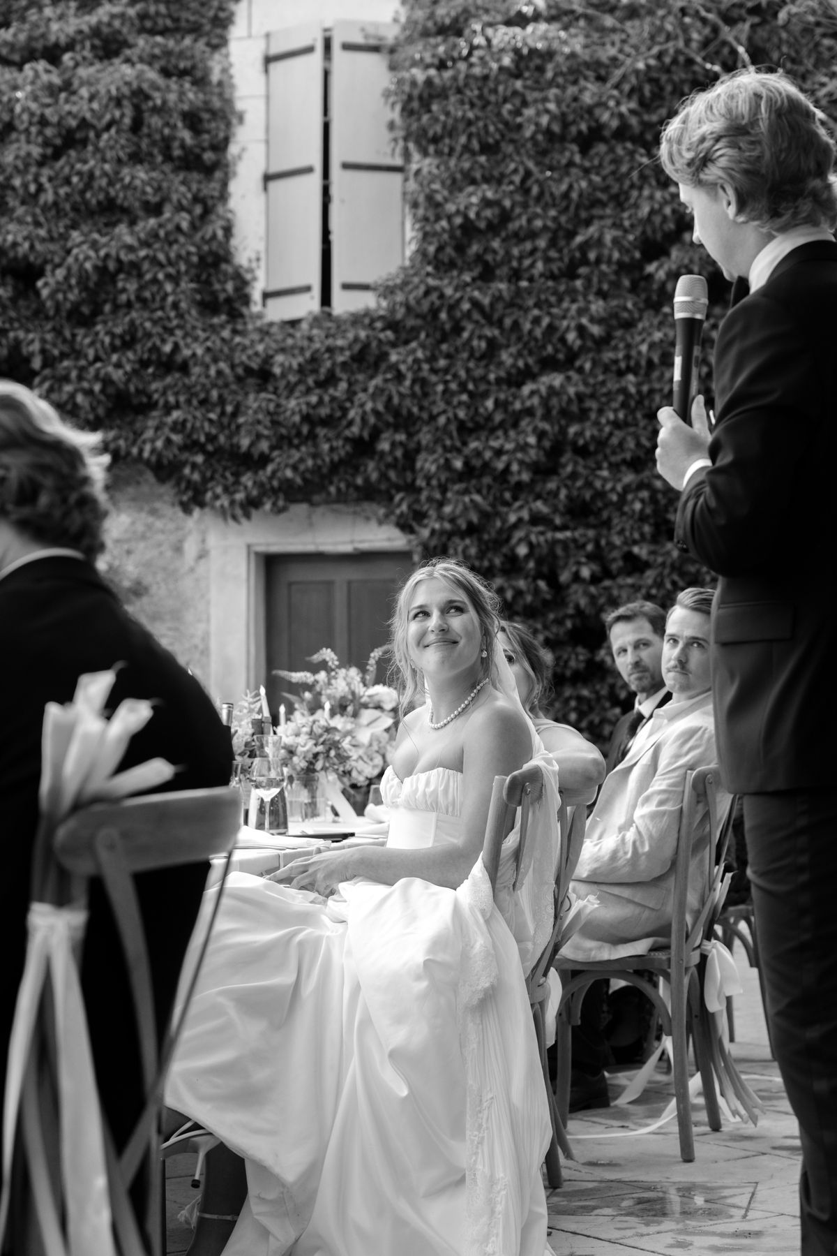 In black and white, a man in a dark suit speaks into a microphone while the bride in a gown laughs at a table.