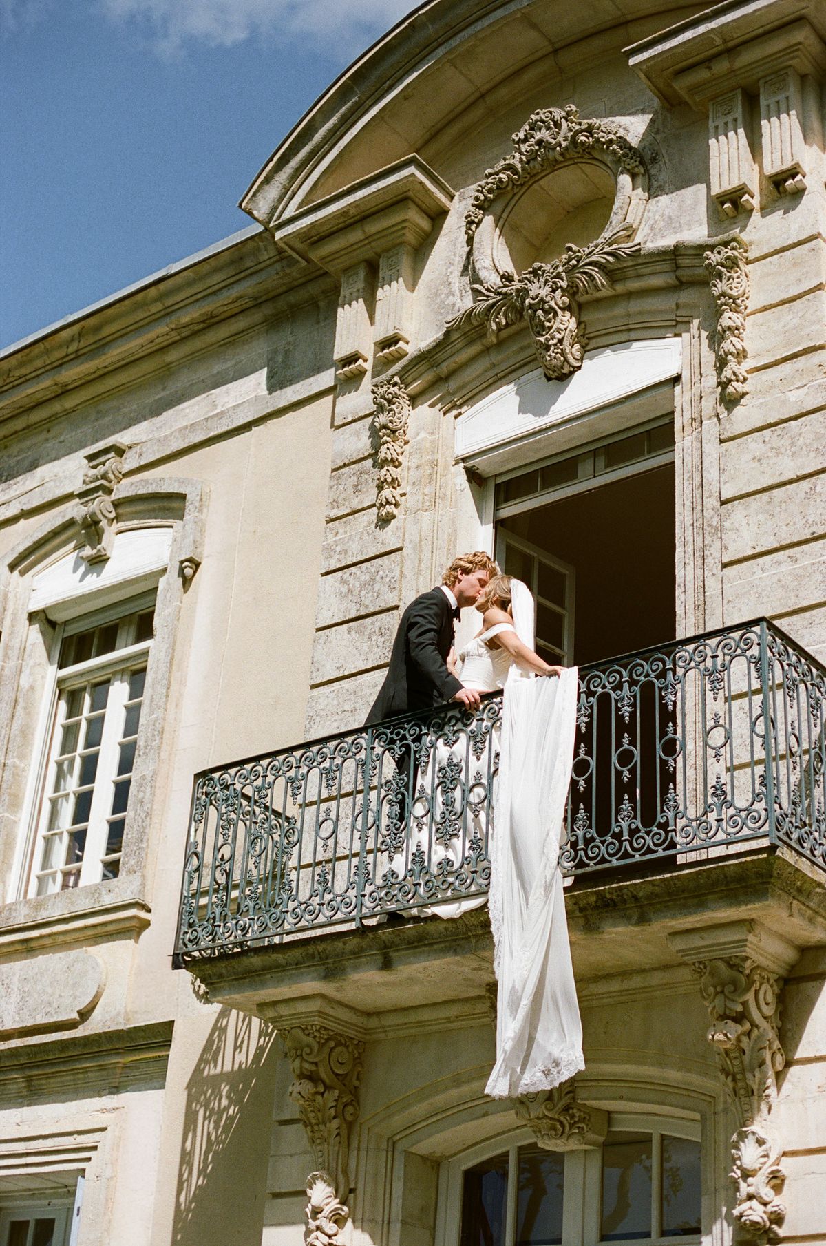 The couple stand on a wrought-iron balcony of a stone chateau while the bride's long train hangs below.