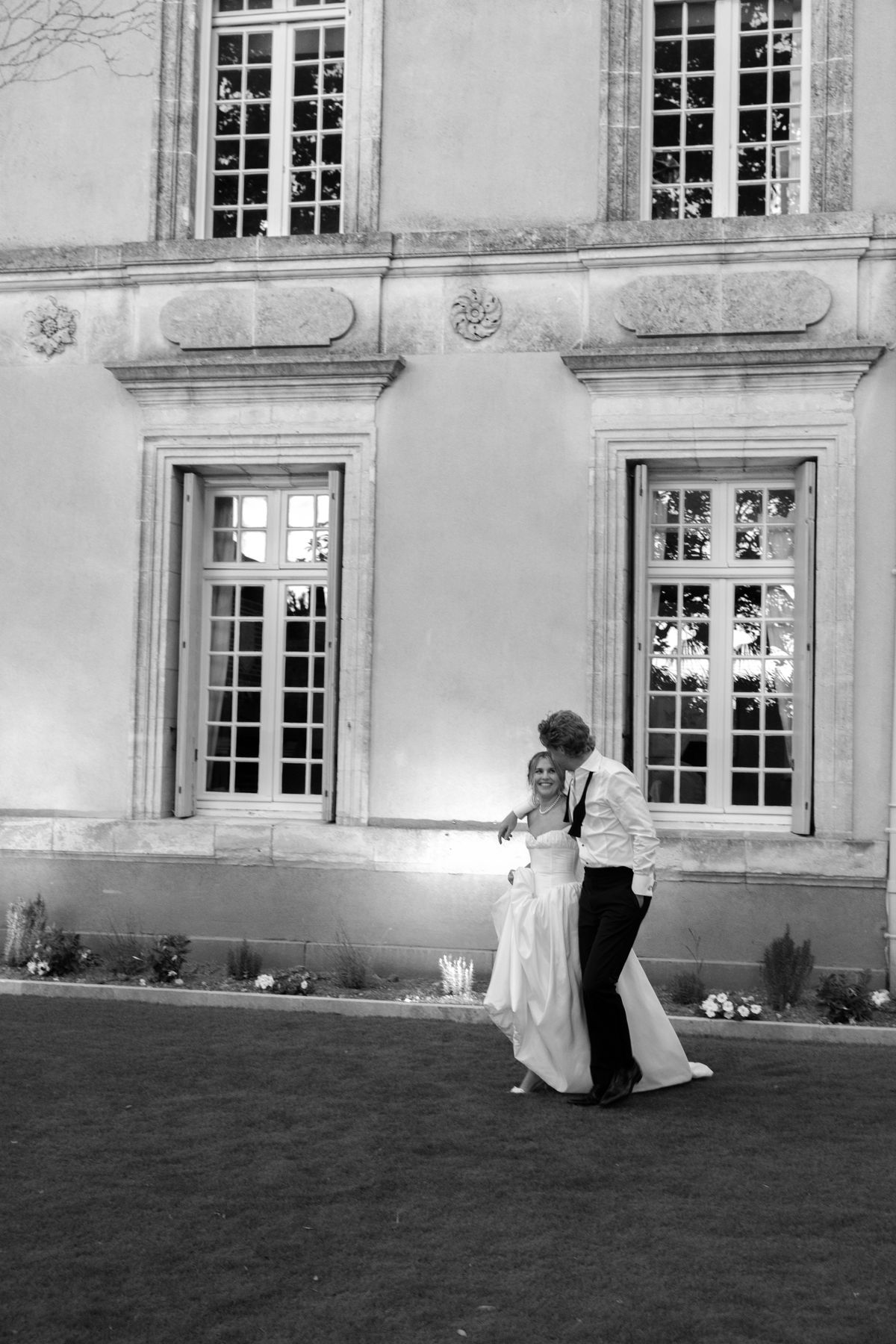 In black and white, the couple embrace and dance on a lawn in front of a stone chateau with tall windows.