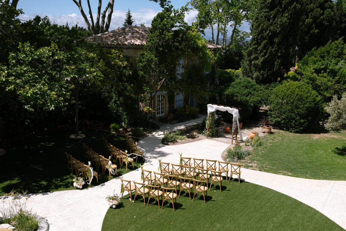A drone view of wooden chairs on a lawn beside a curving white path leading to a floral arch, with a stone house behind.