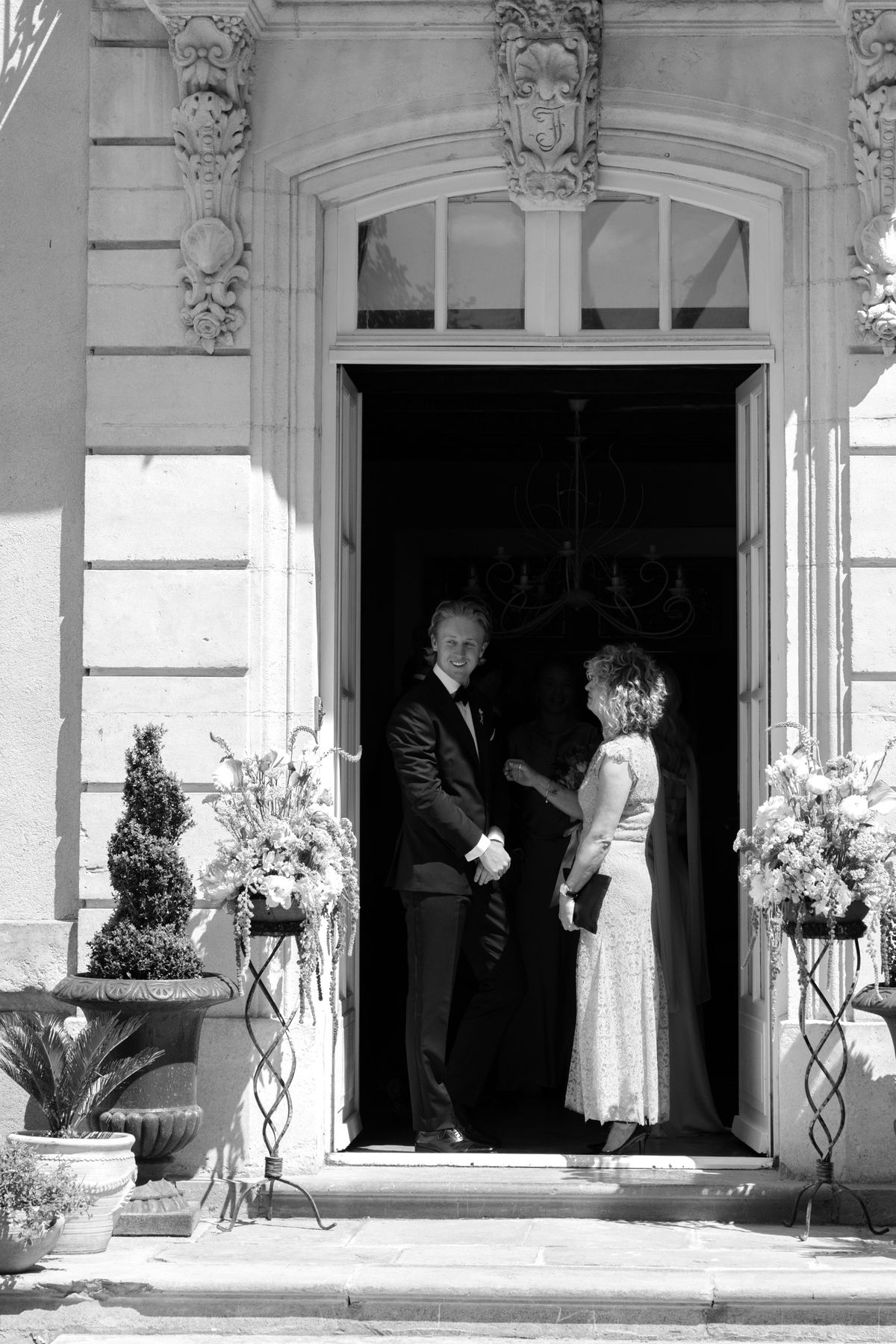 A smiling groom in a dark suit and bow tie talks with a woman guest in a lace dress in a grand stone doorway.