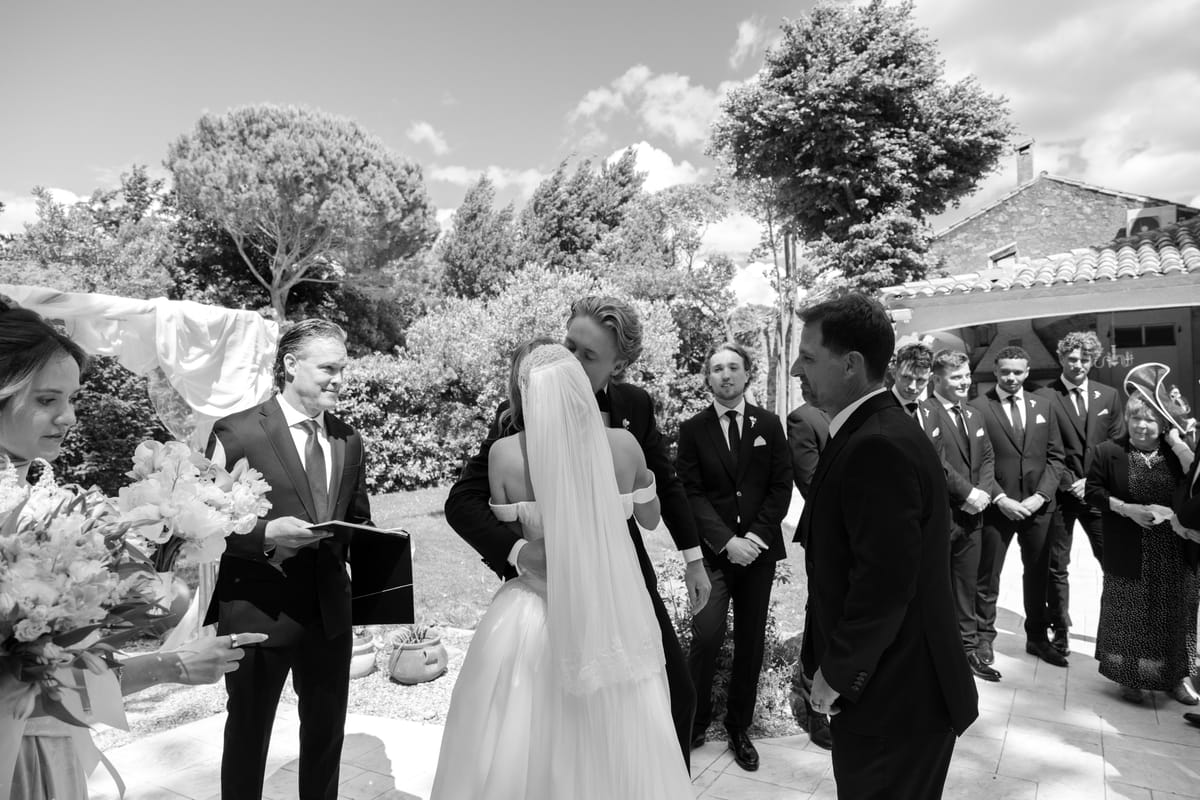 In black and white, the bride in a long veil stands before an officiant and men in dark suits in a garden.