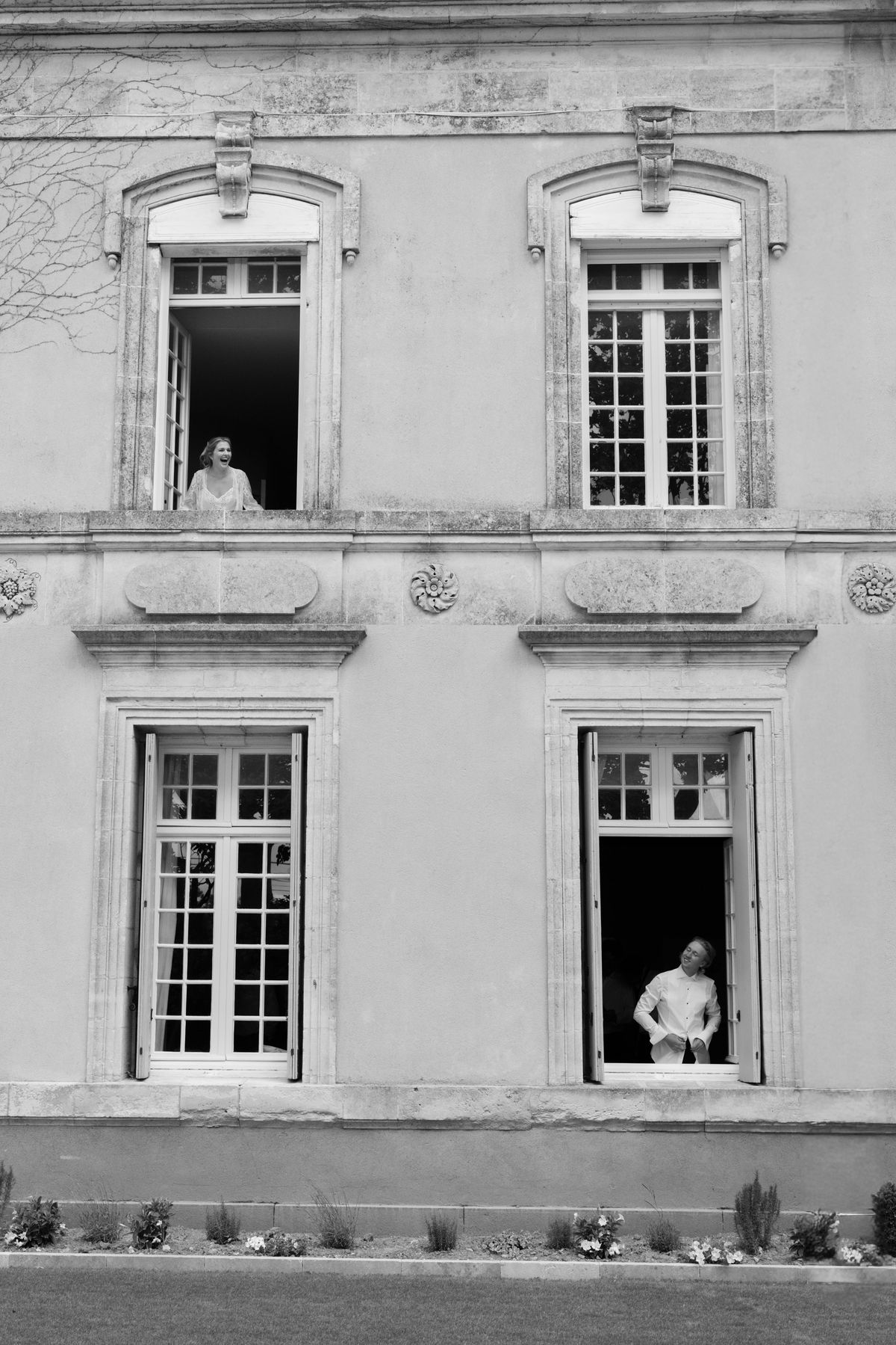 Two people lean from separate windows of a grand stone house: a woman in an upper window, a person in white below.