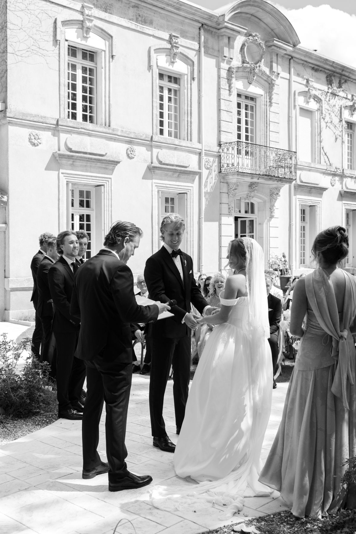 The bride and groom hold hands facing each other at the outdoor ceremony in front of the château, in black and white.