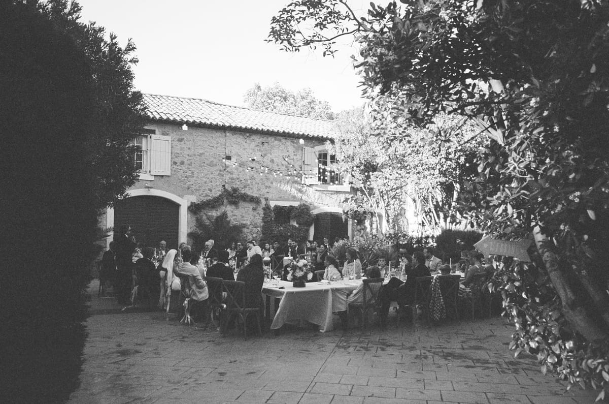 In black and white, guests sit at long dinner tables in a paved courtyard beside a stone building and trees.