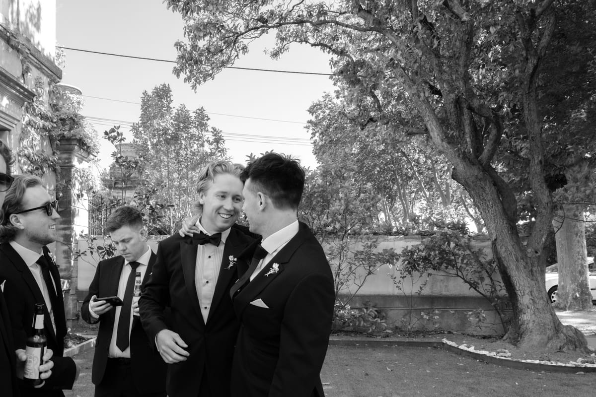 Groomsmen in black tie share a hug and a laugh outside before the ceremony, in black and white.