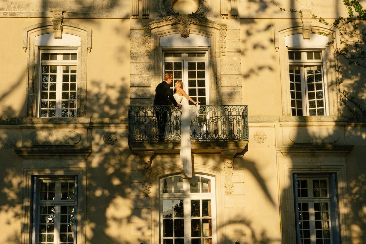 A couple embrace on an ornate wrought-iron balcony of a golden stone mansion, the bride's long white dress hanging below.