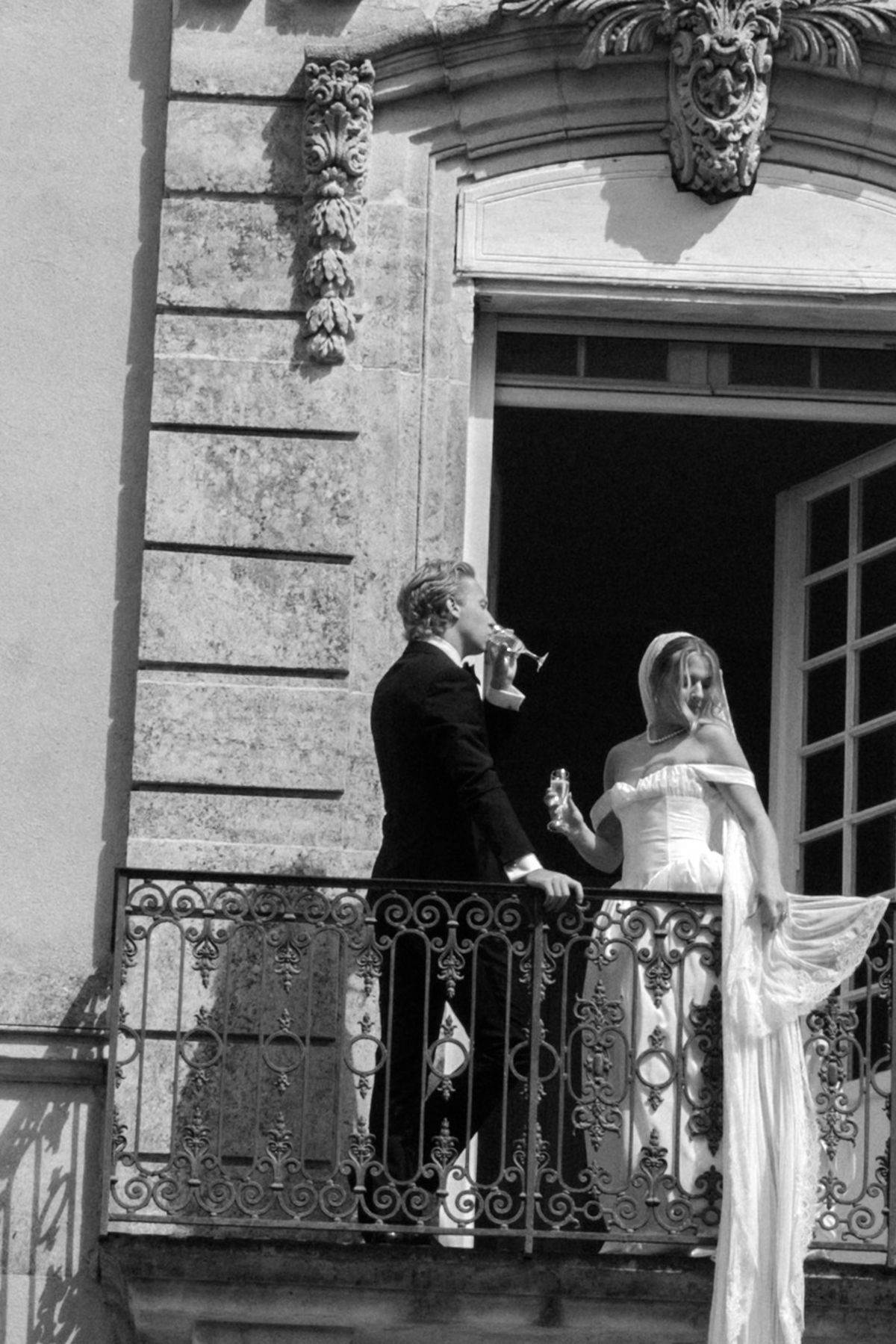 In black and white, the groom and the bride in a long gown stand together on a balcony with an ornate iron railing.