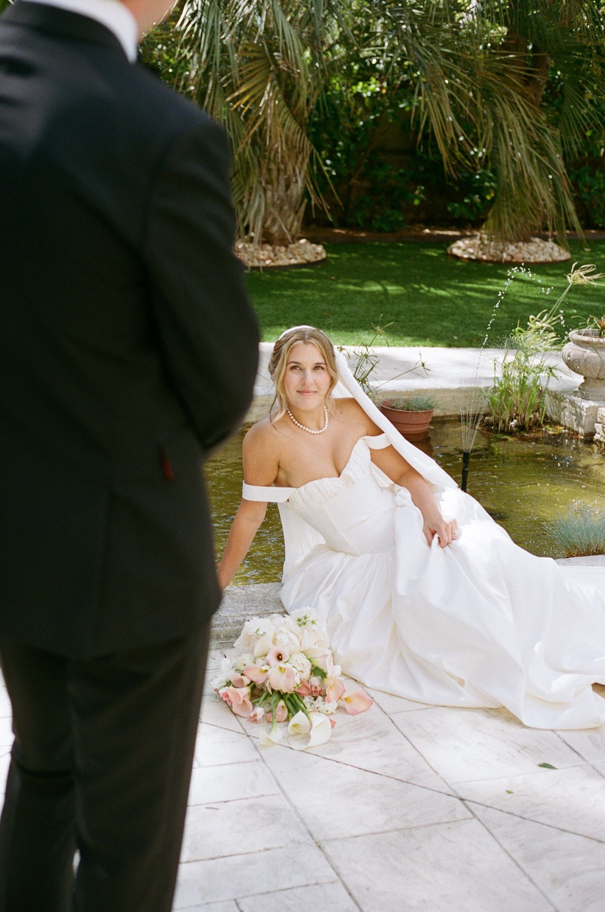 The bride sits on a paved edge beside a pond with a bouquet, while the groom stands in the foreground with his back turned.
