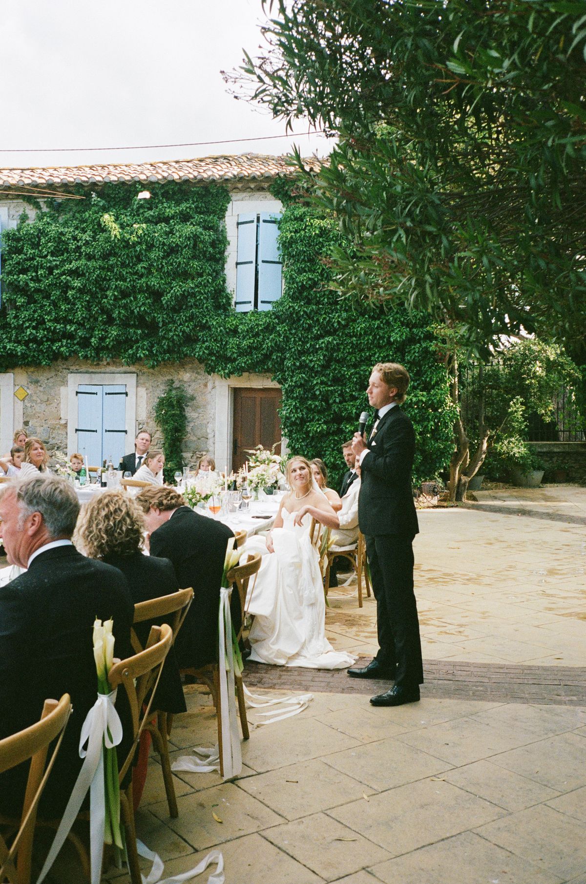 In a courtyard, the groom stands to speak while the bride and guests sit at a long table beside an ivy-covered building.