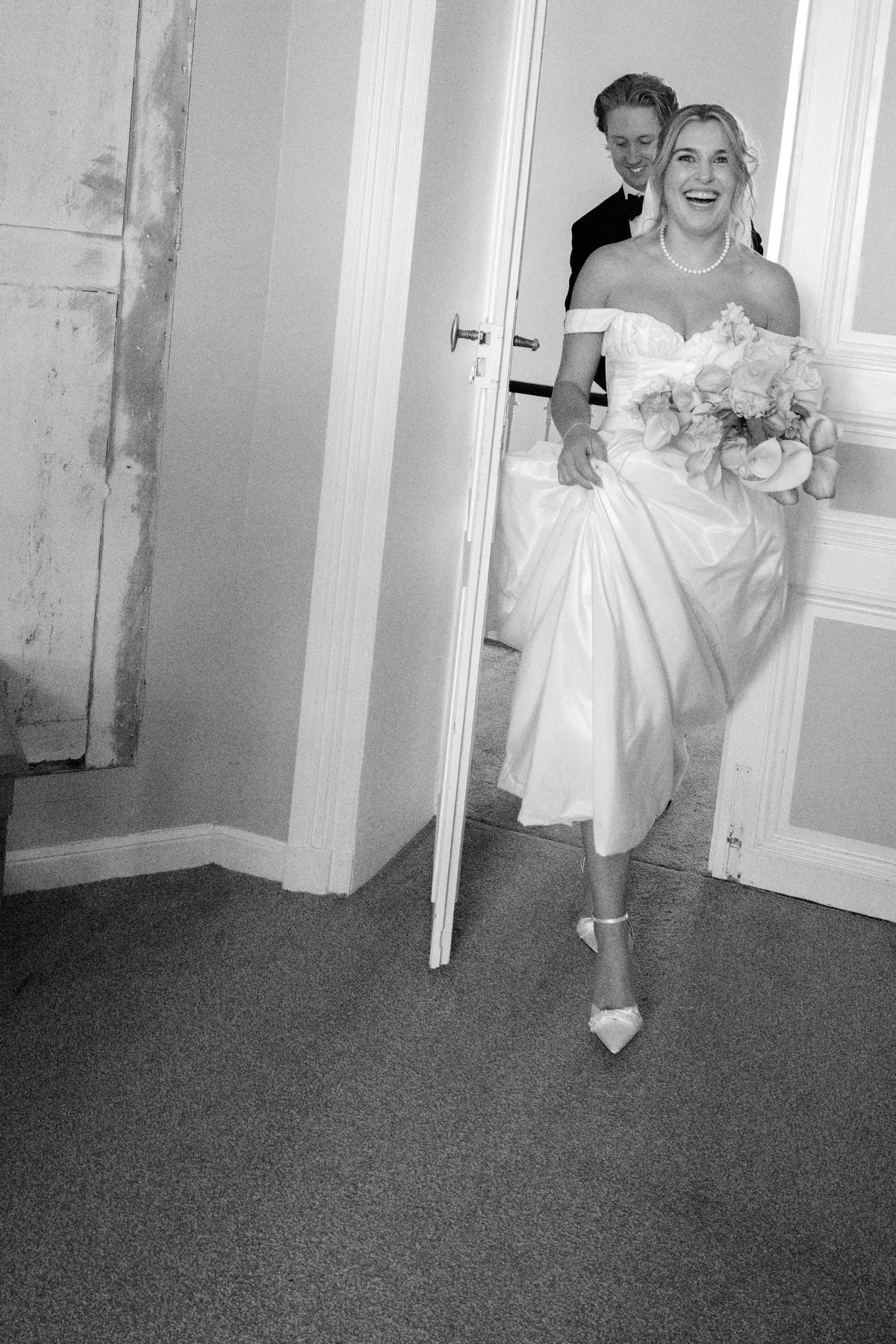 In black and white, the laughing bride lifts her gown and steps through a doorway, with the groom smiling behind her.