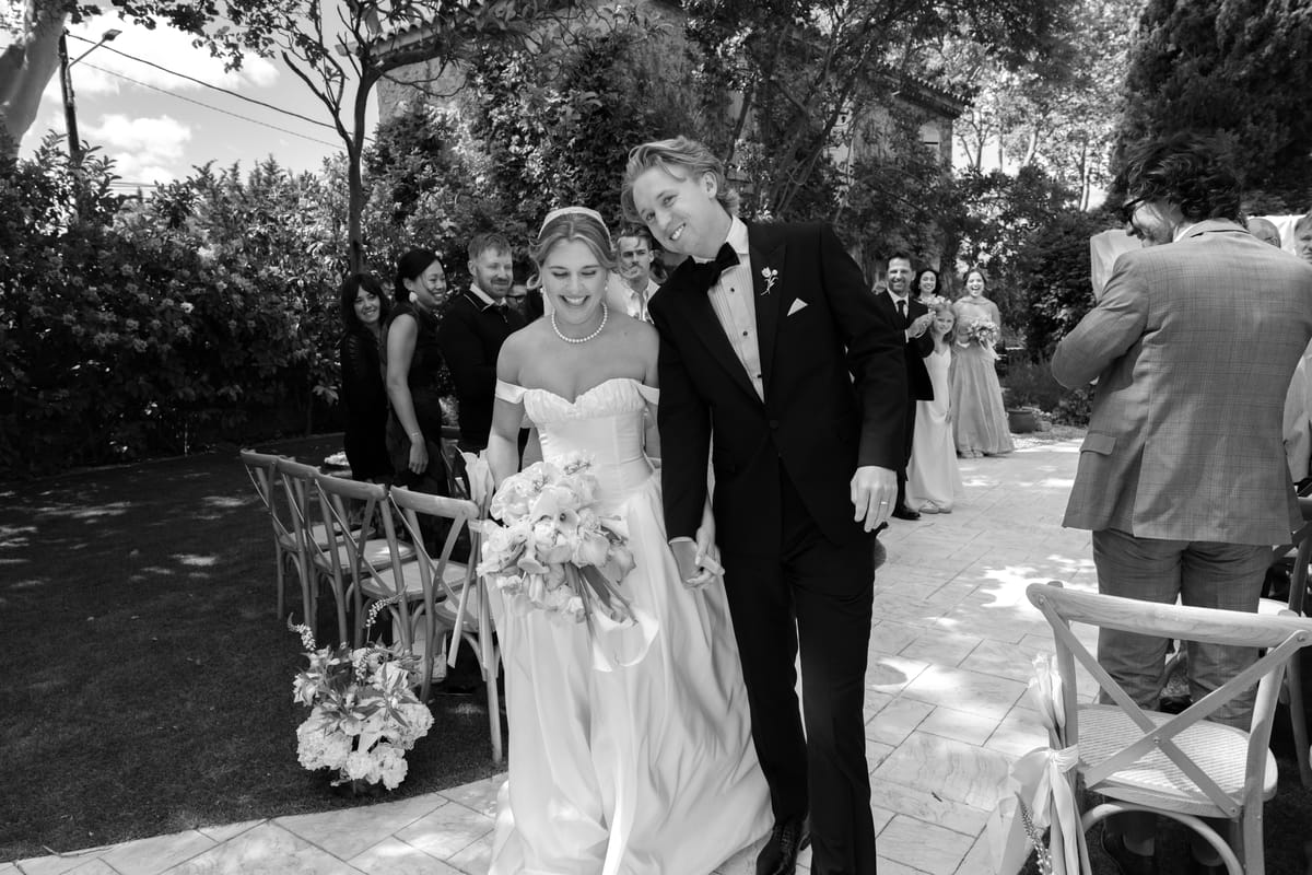 The bride holding a bouquet and the groom walk along a path between ceremony chairs, with guests smiling around them.