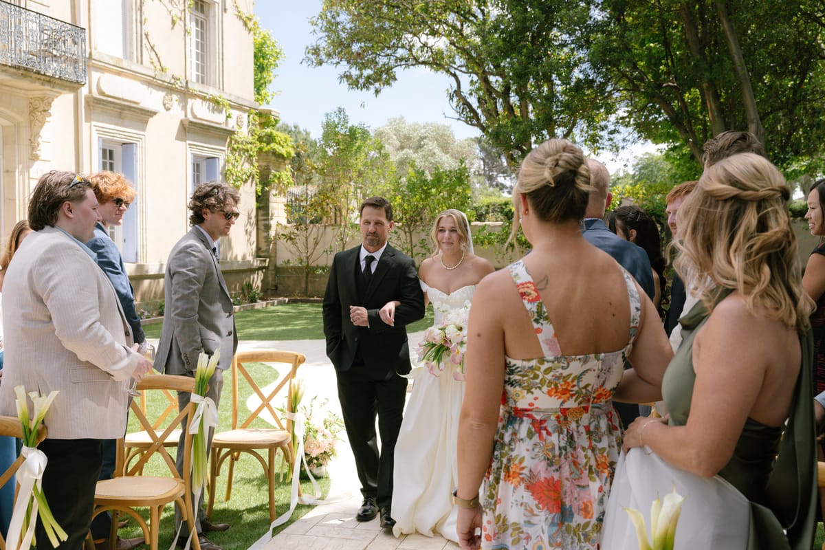 The bride walks arm in arm with a man in a dark suit past seated guests in a sunny garden, with wooden chairs along the aisle.