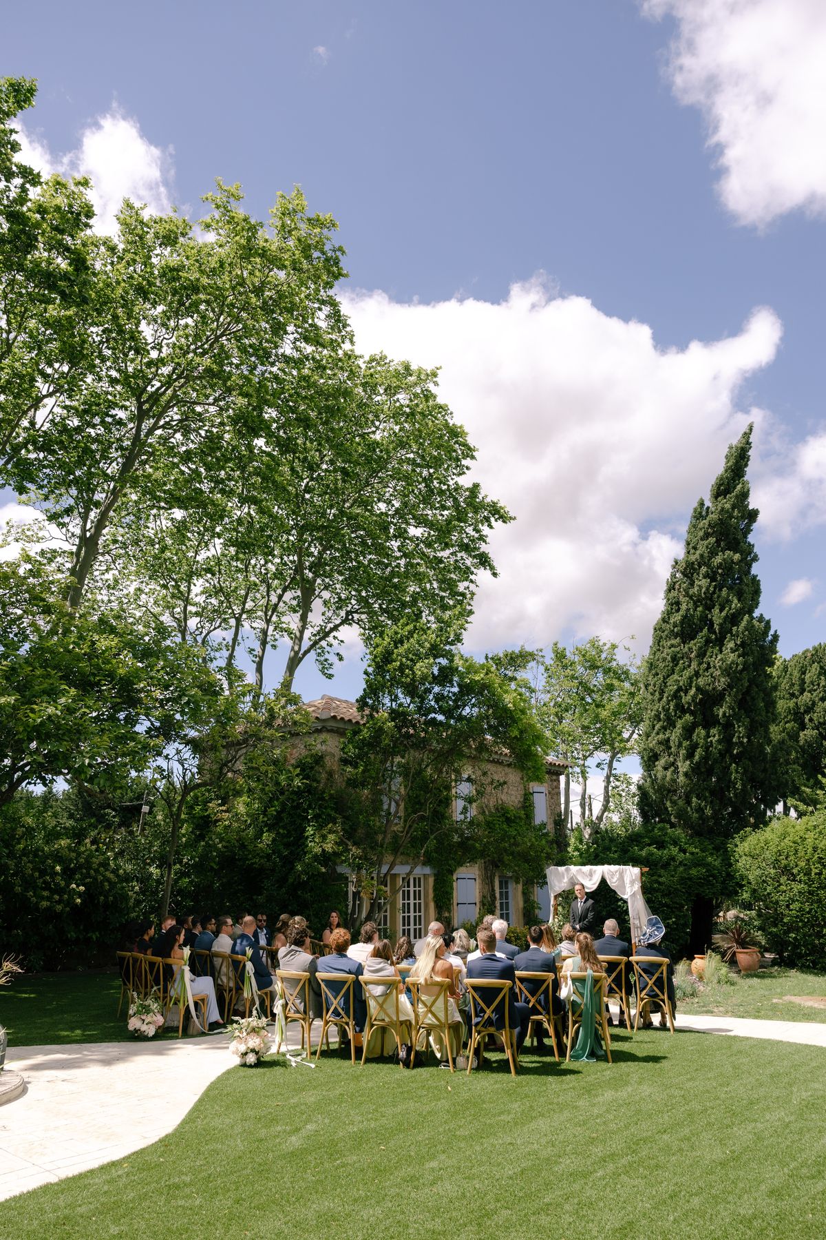 Seated guests face a person standing by a white draped canopy during an outdoor ceremony on a lawn beneath tall trees.