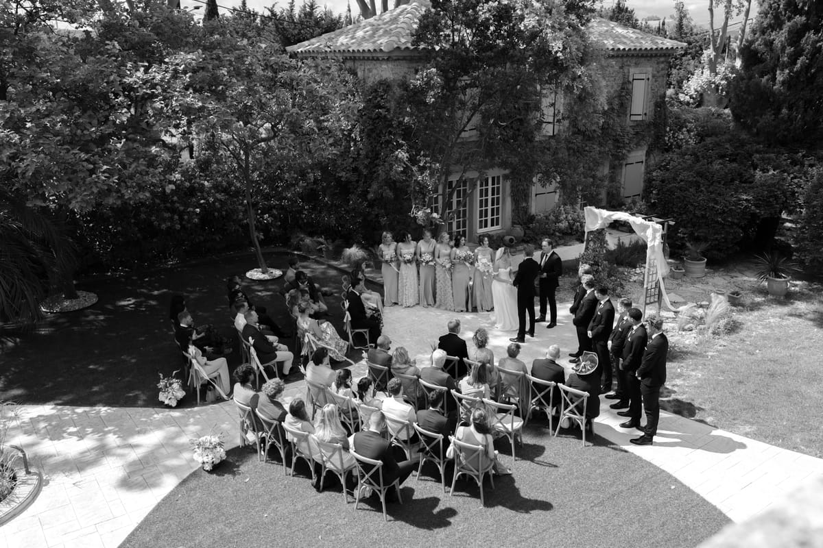 In black and white, seen from above, guests sit on chairs facing the couple and wedding party beneath an arch in a leafy garden.