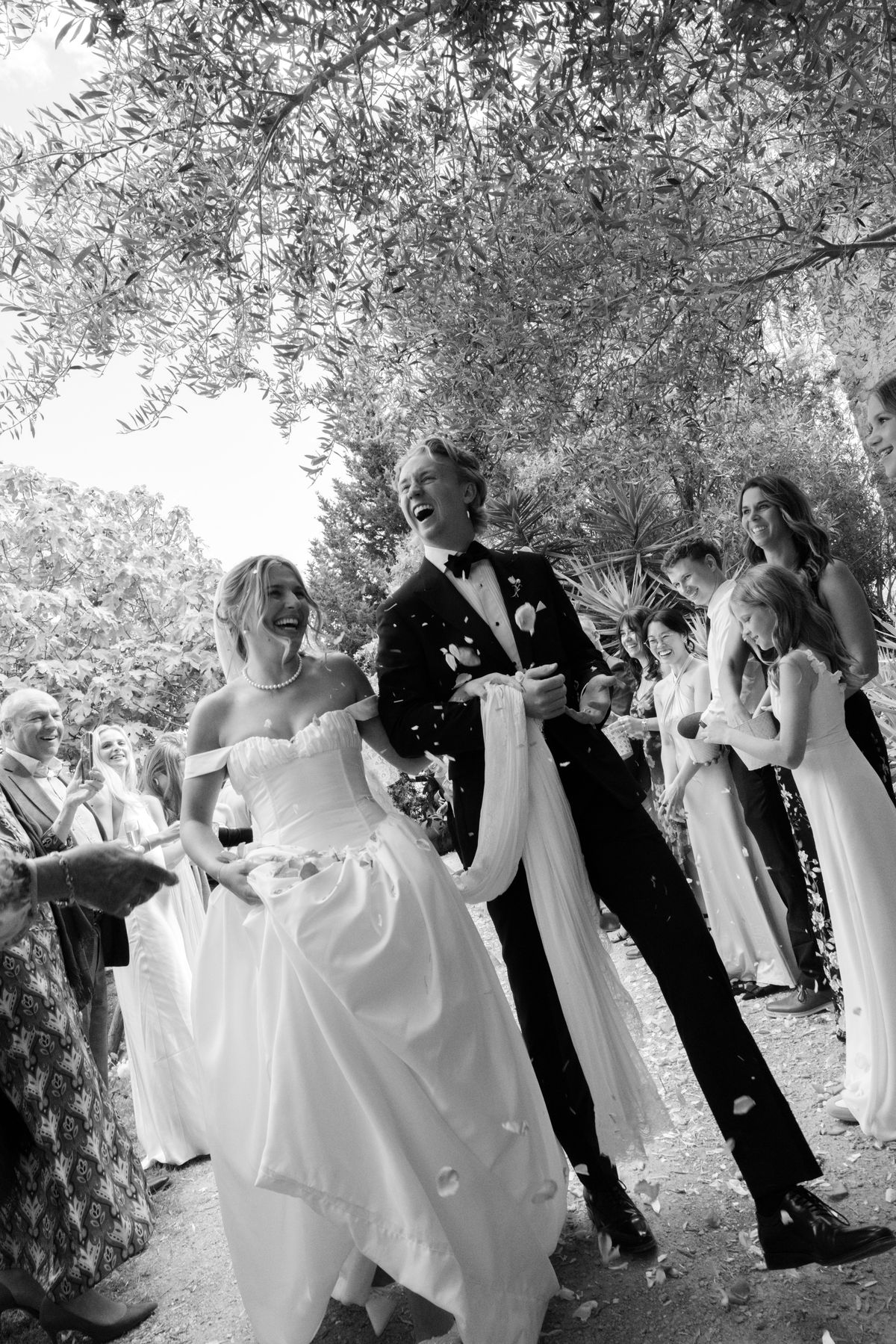 In black and white, the bride and groom walk laughing beneath trees, the bride holding her skirt and a long ribbon, with guests around them.