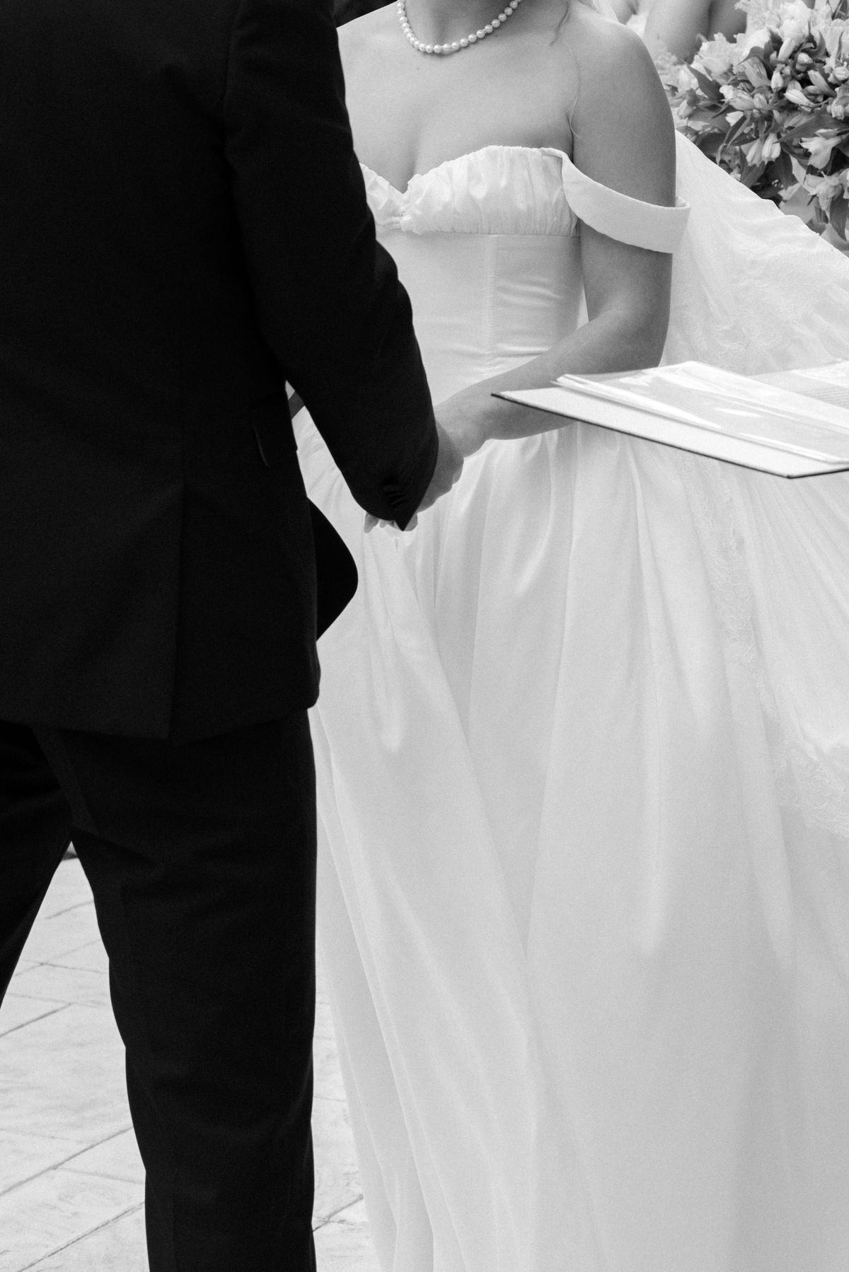 The bride in a strapless gown with a pearl necklace holds a folded paper while the groom in a dark suit holds her hand.