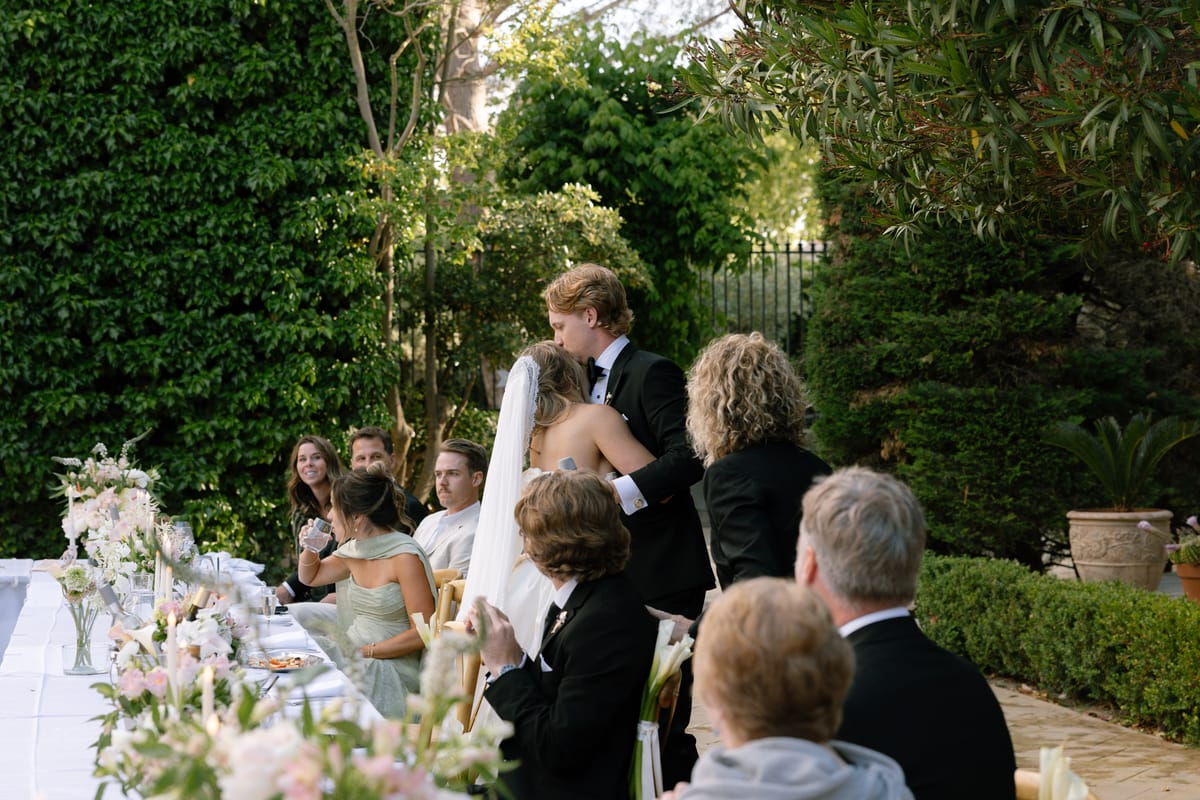 The couple embrace beside a long outdoor table decorated with pink flowers while seated guests watch and smile.