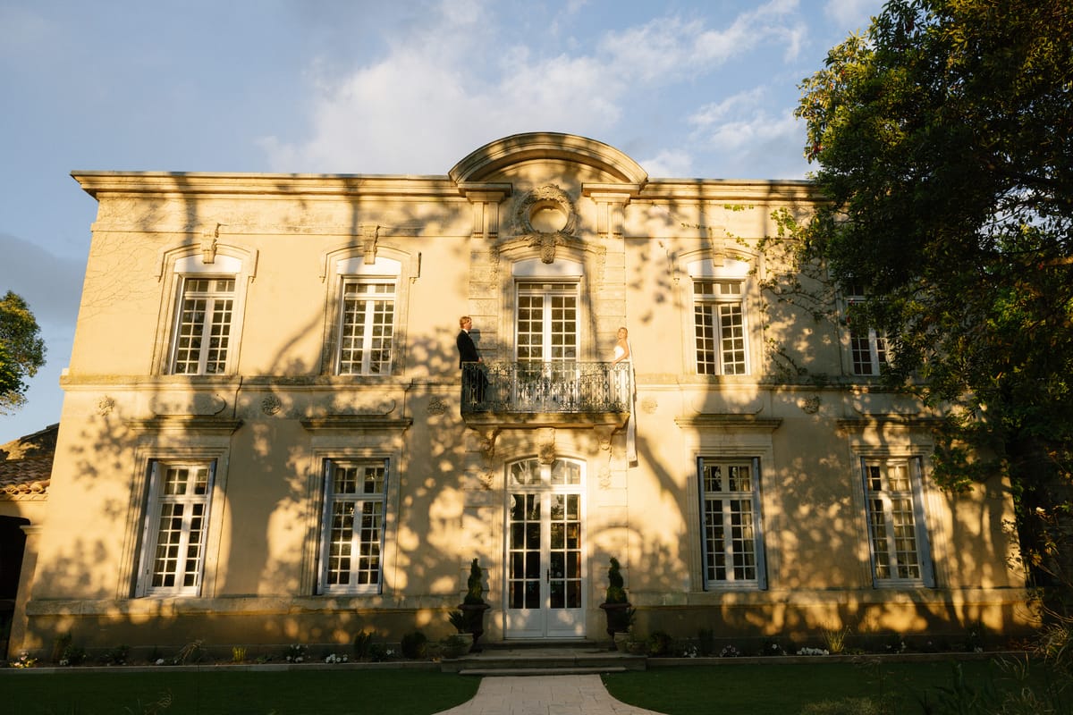 Two people, apparently the couple, stand on a small balcony of a grand cream stone château lit by warm evening light.