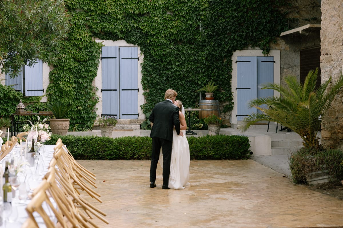 The couple embrace in a stone courtyard beside a long dressed table, with ivy-covered walls and blue shutters behind.