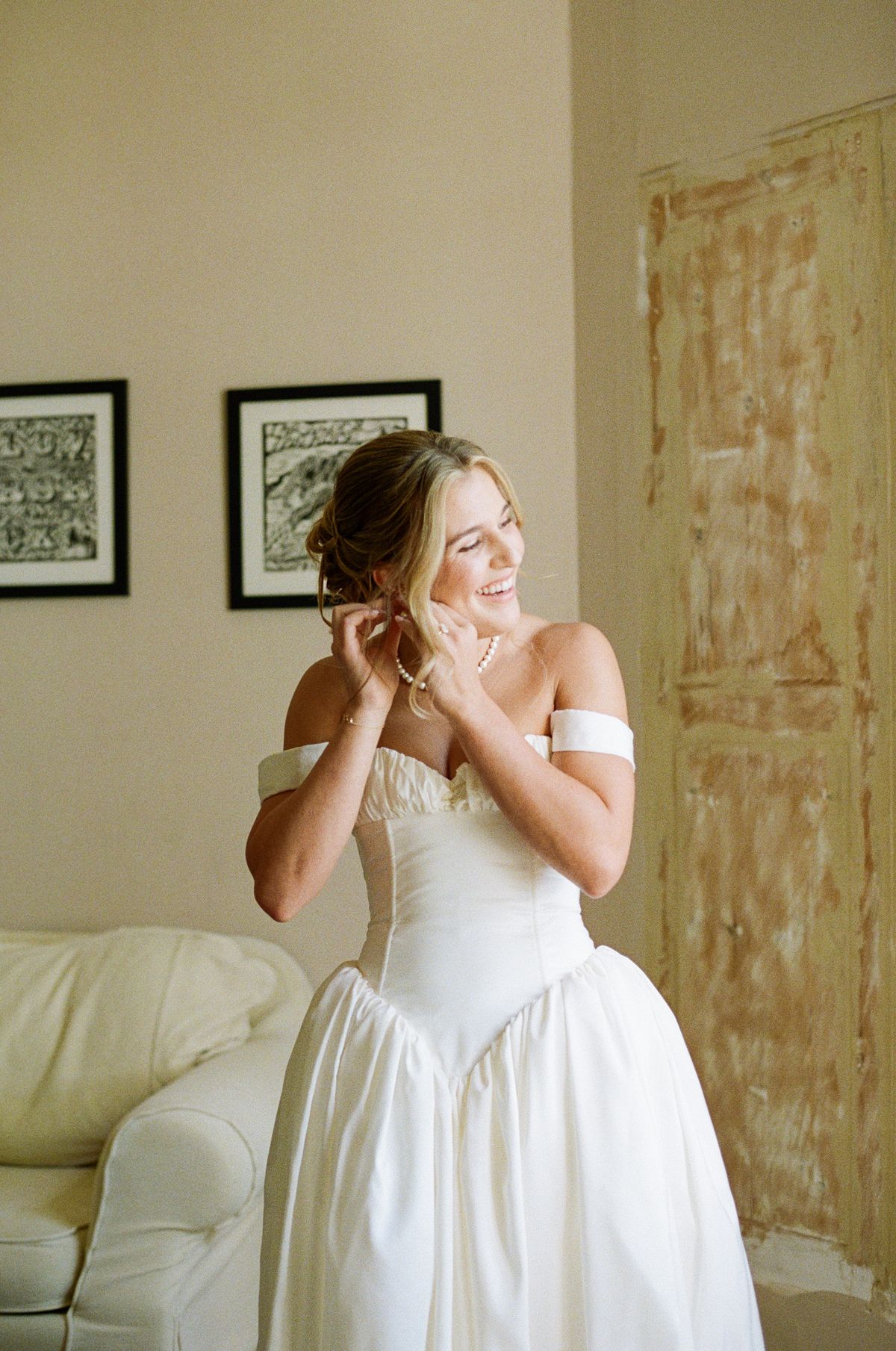 The bride in an off-the-shoulder white gown smiles to one side as she fastens an earring in a softly lit room.