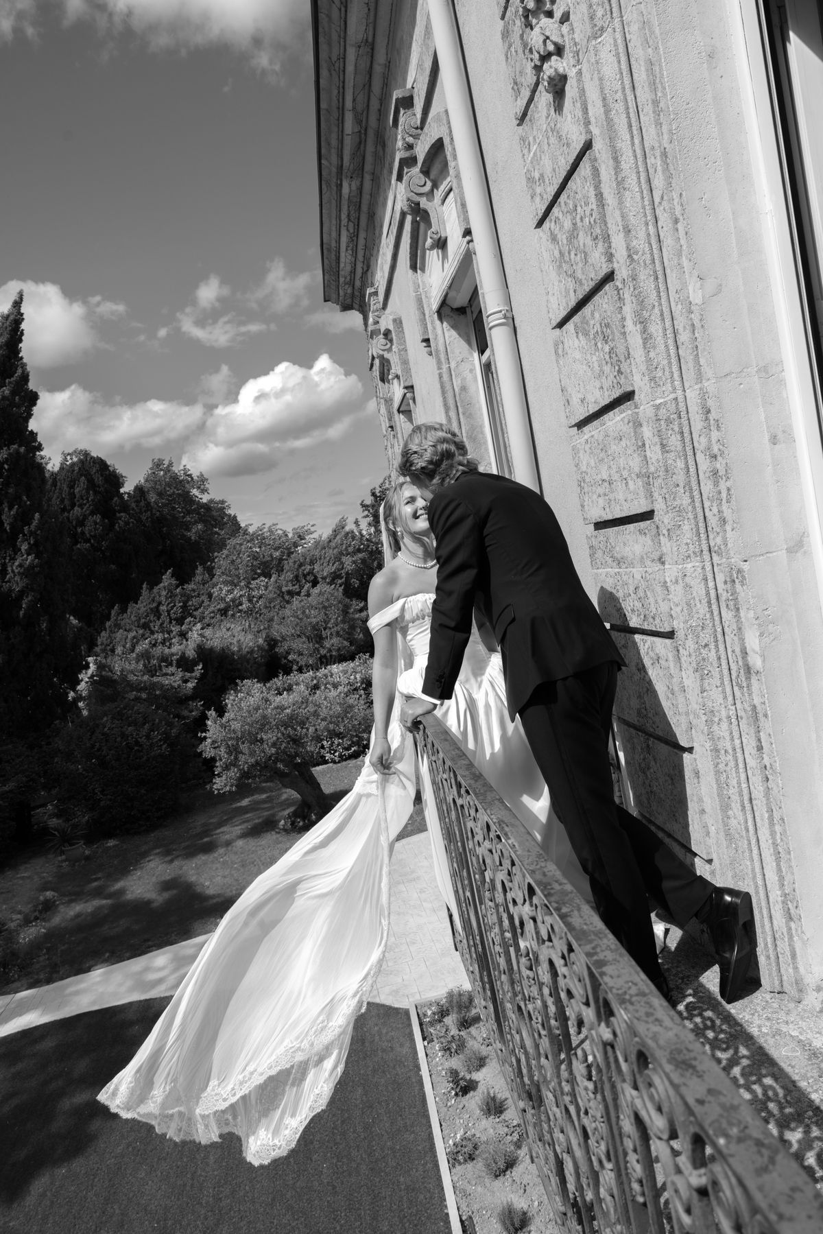 The groom leans over an ornate balcony railing beside the bride, whose long white skirt flows in the wind, beside a stone facade.