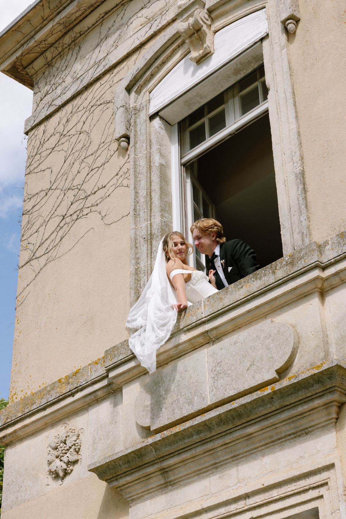 The bride in a veil and the groom lean out of an open upstairs window of a stone chateau, smiling at each other.