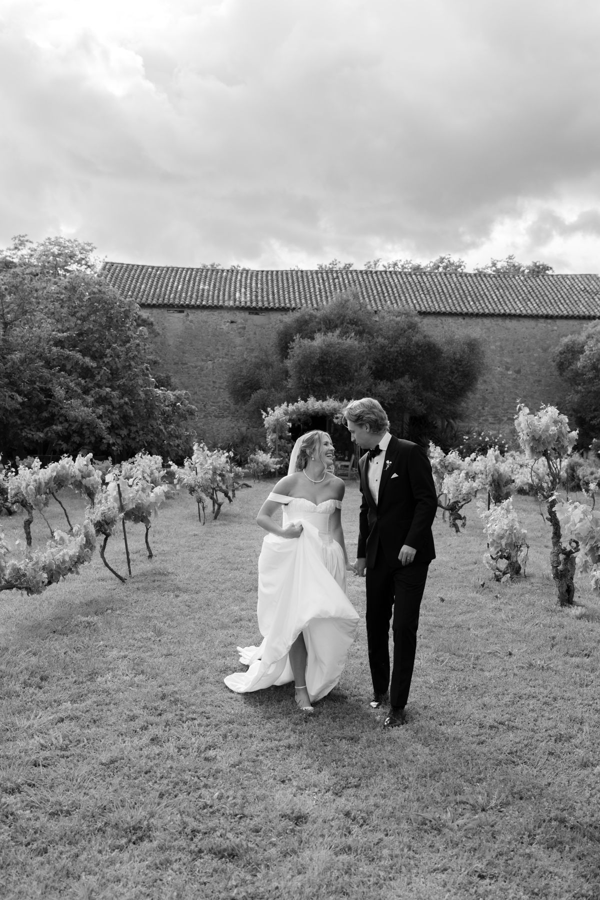In black and white, the couple walk side by side across grass between vines, with a stone wall and cloudy sky behind.