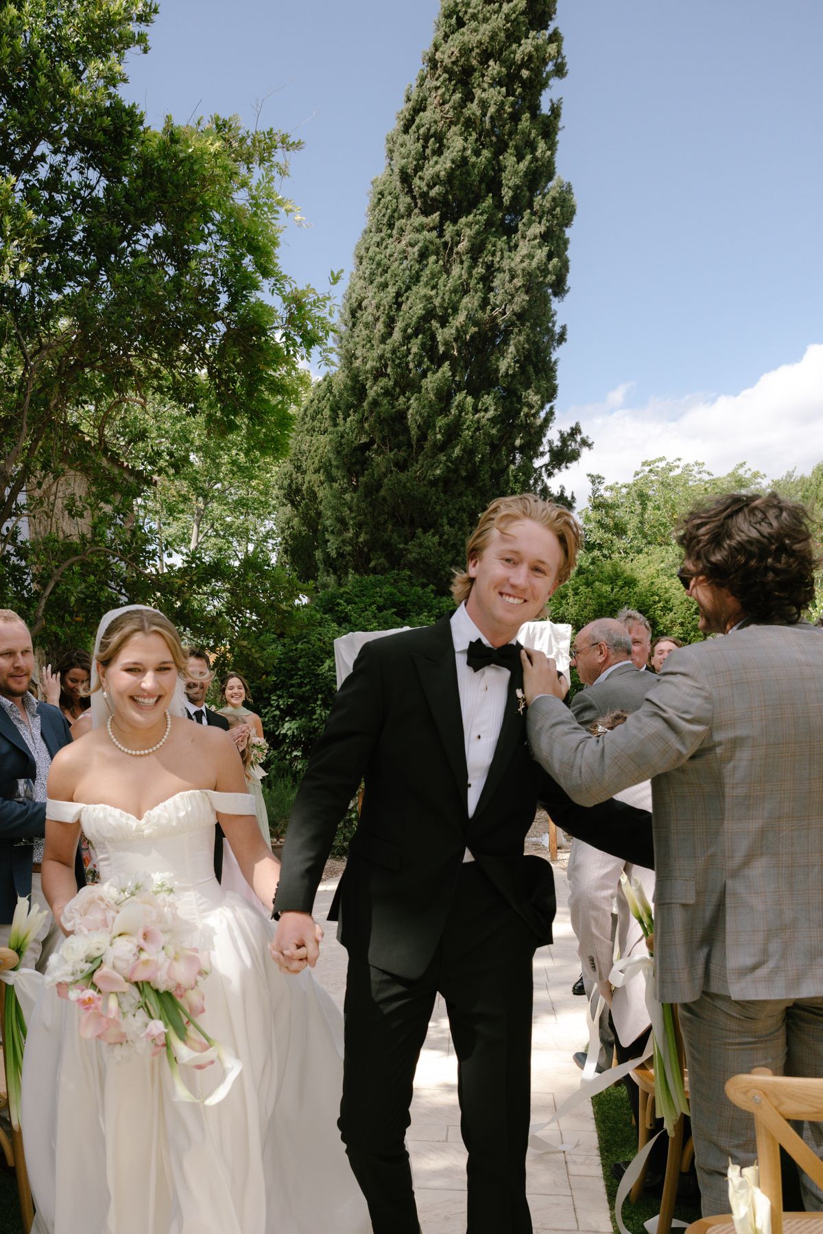 The smiling couple walk hand in hand through a garden aisle while a guest pats the groom on the shoulder.
