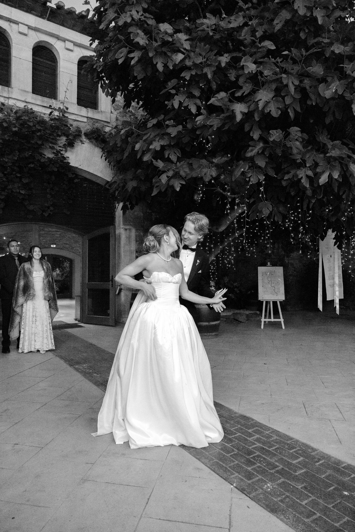 In black and white, the bride in a full-skirted gown stands in a courtyard with the groom close behind and a guest in the distance.