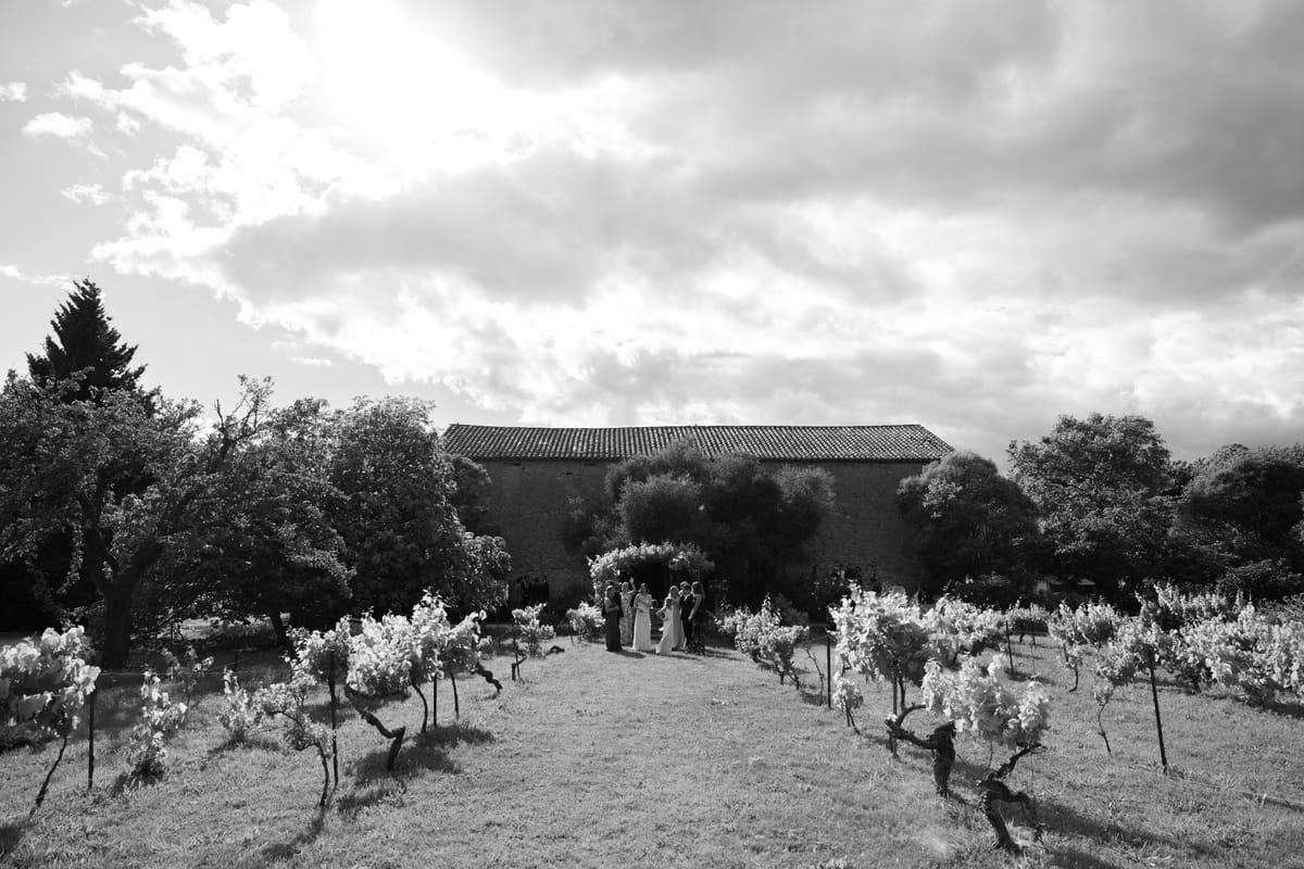 A small group of guests stands far off between rows of vines, before a long stone barn under a dramatic cloudy sky.