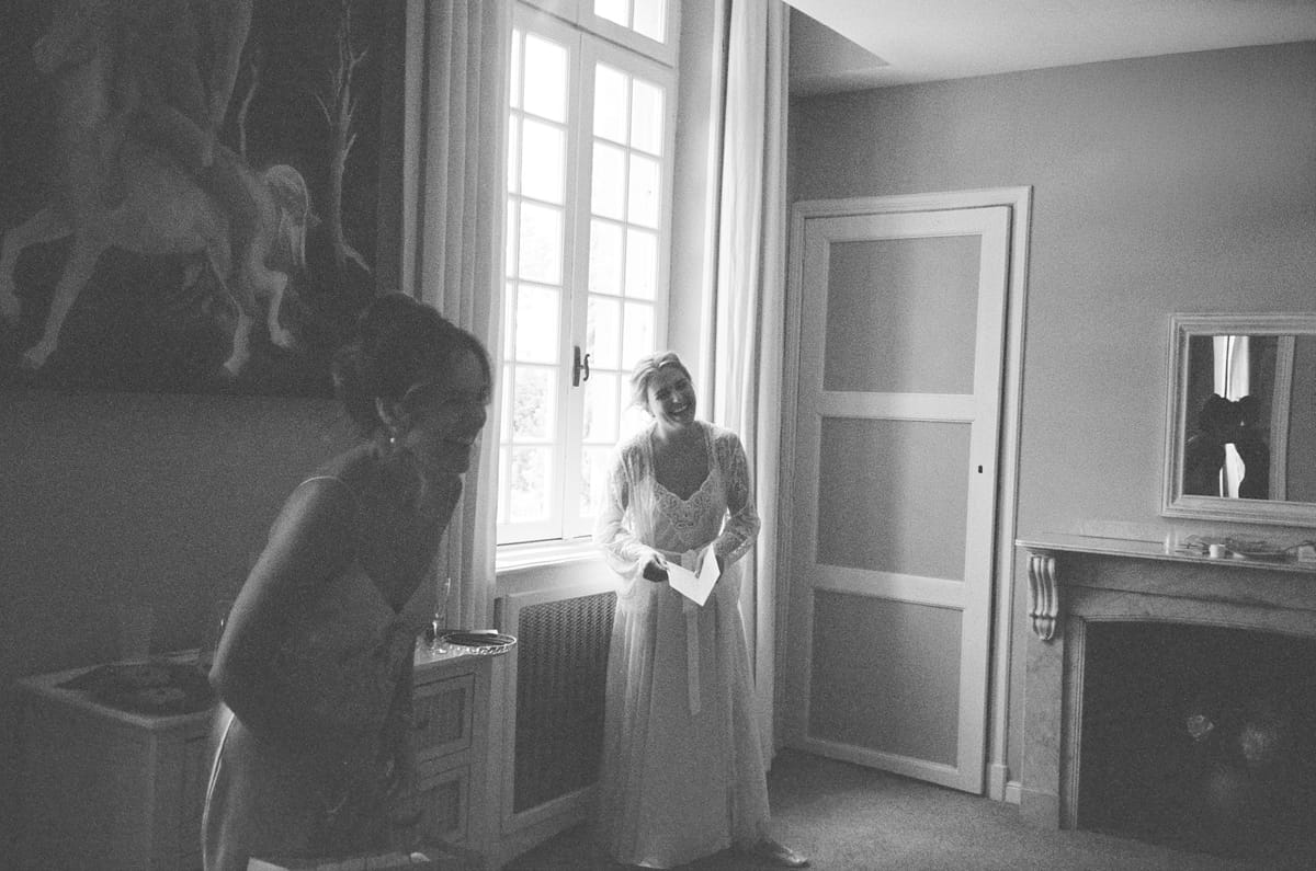 In black and white, the bride stands beside a tall window in a plain room while another person leans in at the left.