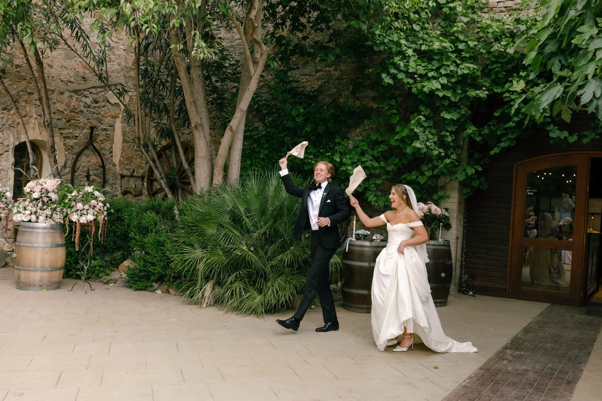 The bride and groom walk across a stone courtyard, each waving a napkin, near flower-topped barrels and green plants.