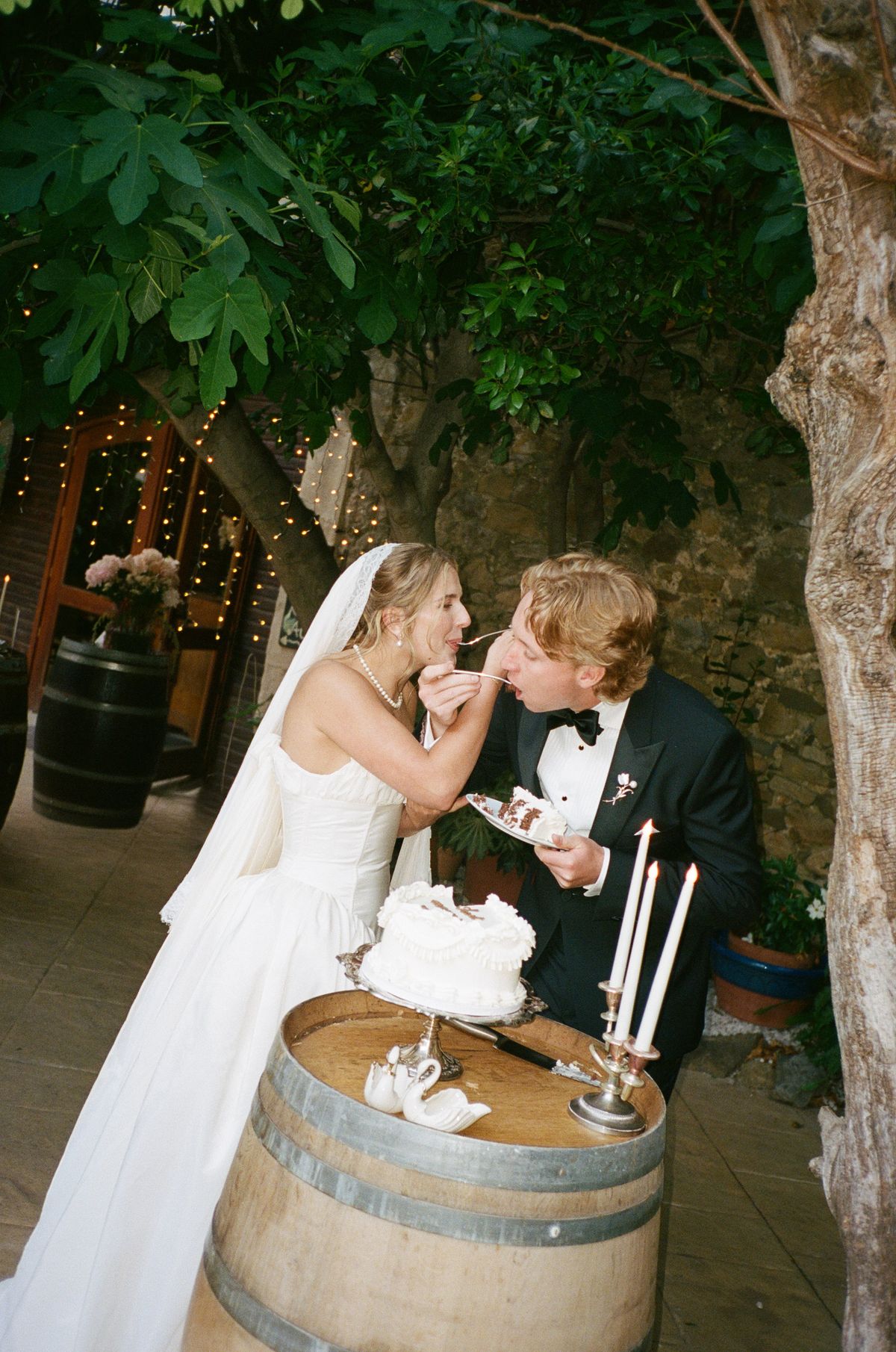 The bride in a veil and the groom playfully feed each other cake beside candles on a wine barrel under a leafy tree.