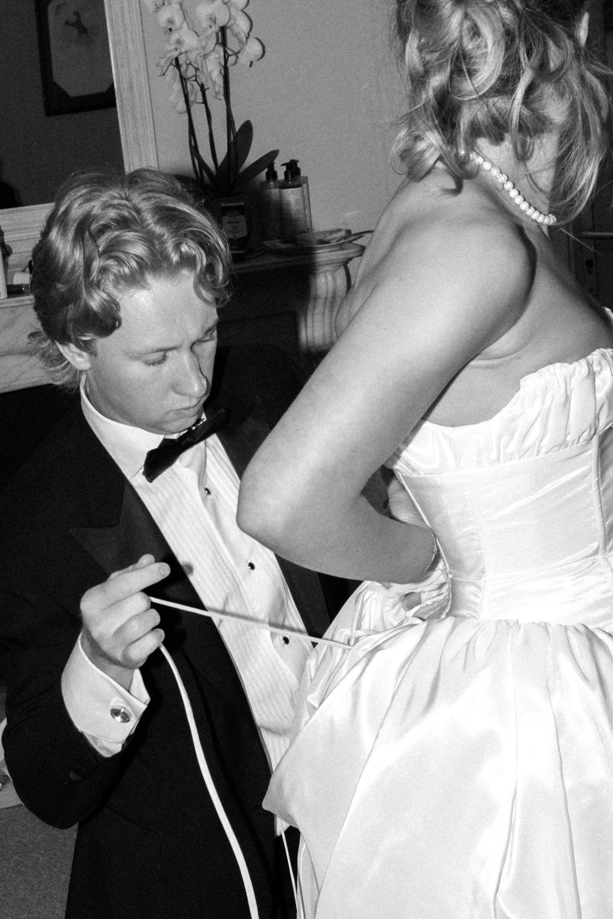In black and white, the groom in a bow tie pulls the lacing at the back of the bride's corseted gown.