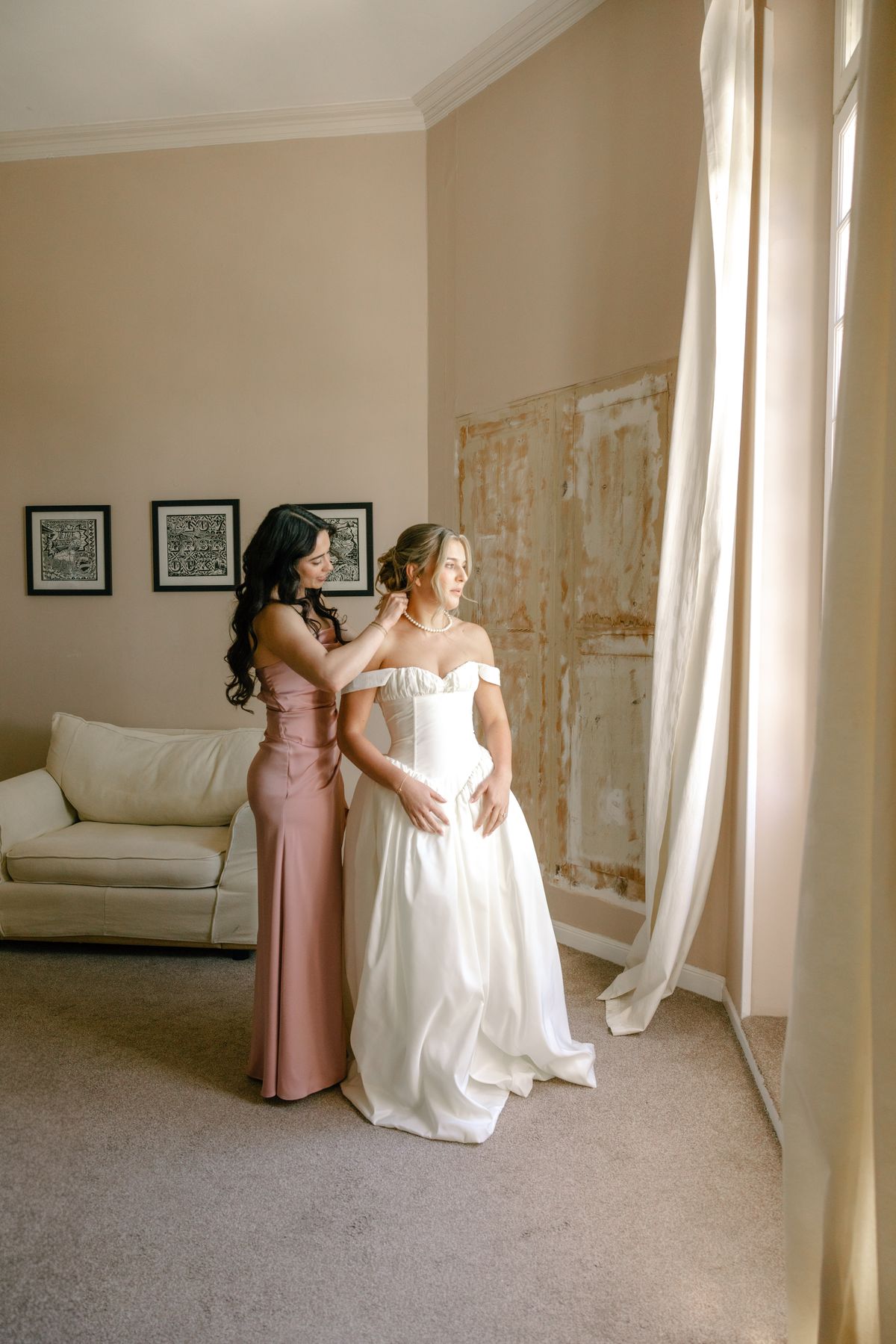 A woman in a long pink dress stands behind the bride in her white gown, both looking towards a window in a softly lit room.