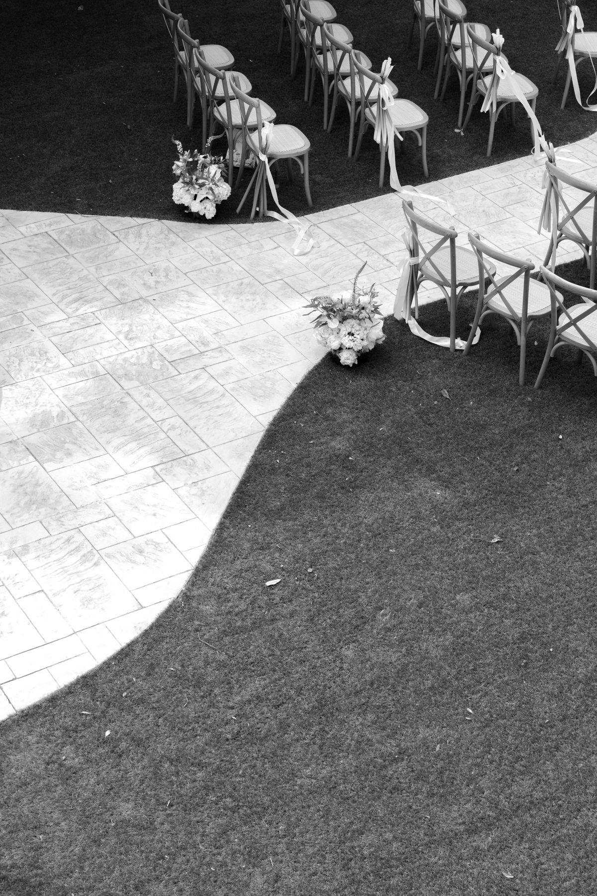 Black and white view from above of wooden chairs with flower arrangements beside a curving paved aisle edged by lawn.